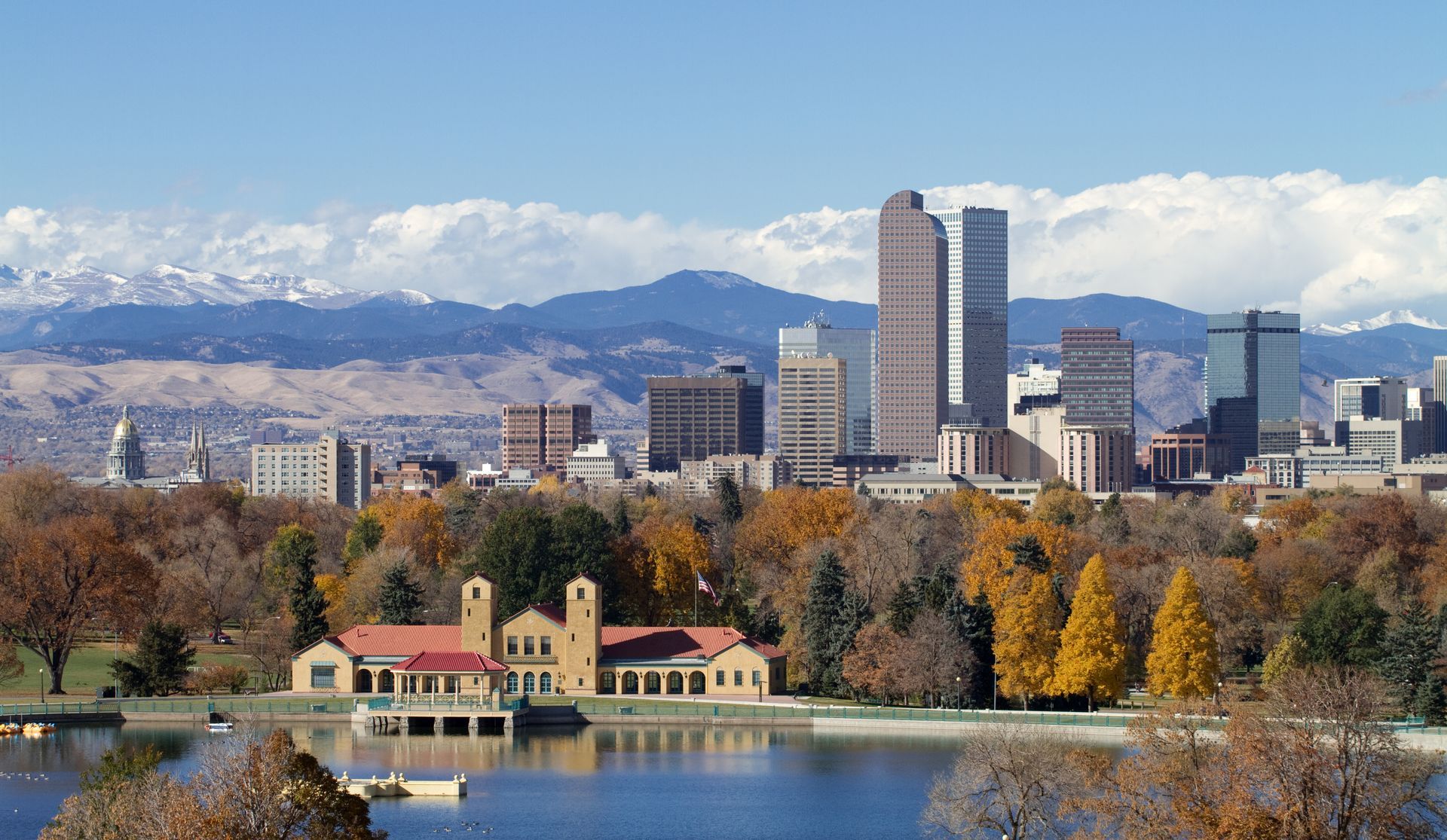 Denver skyline with mountains in the background, a lake in the foreground, and a building with orange trees.