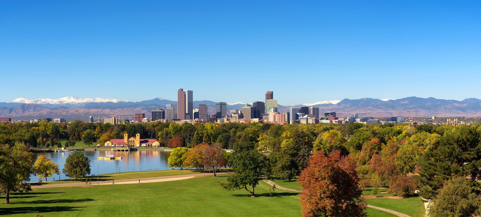 Denver skyline with mountains in the background, a lake, trees, and green grass on a sunny day.
