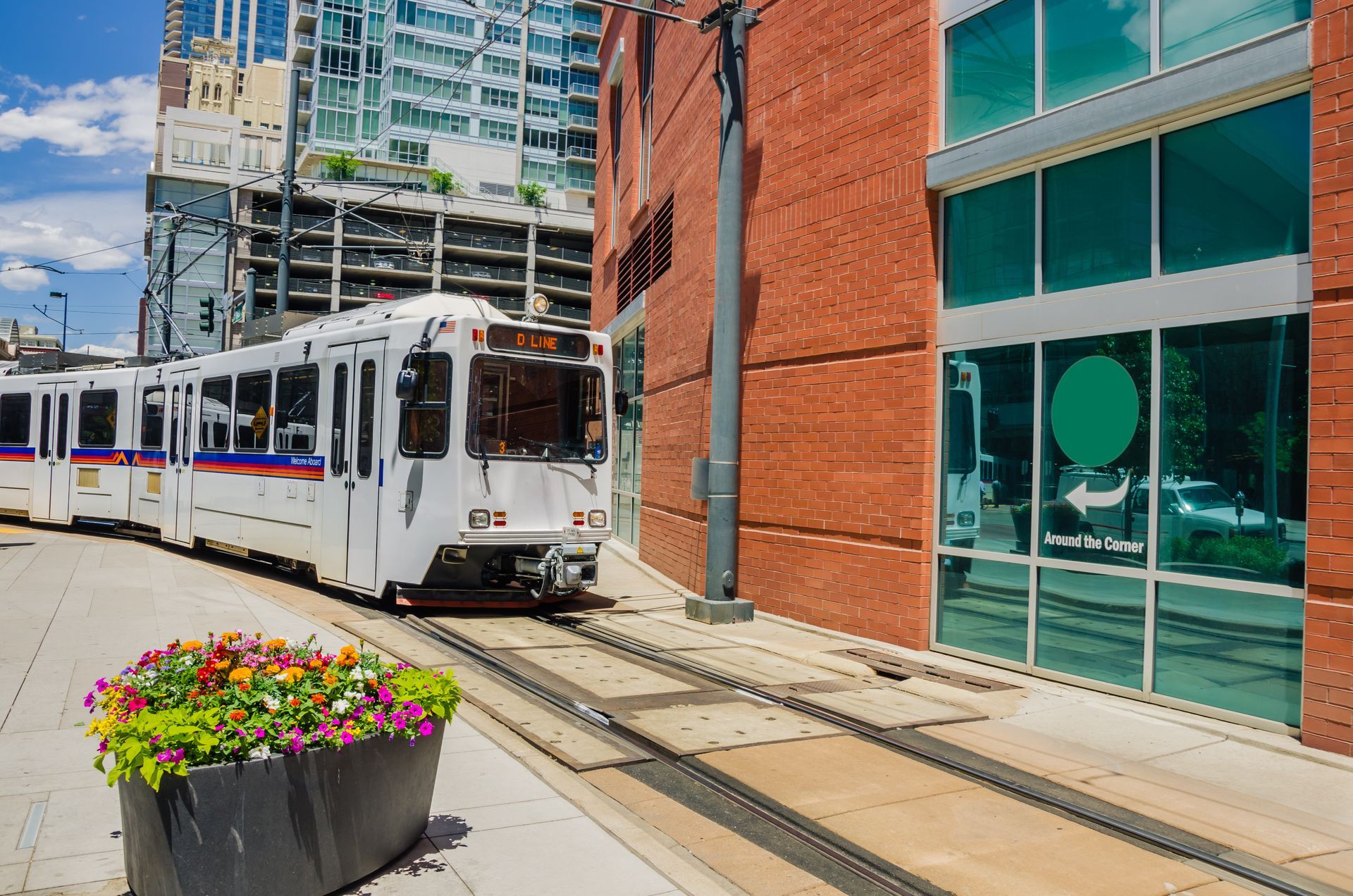Light rail train on tracks next to a brick building and flower box in an urban setting.