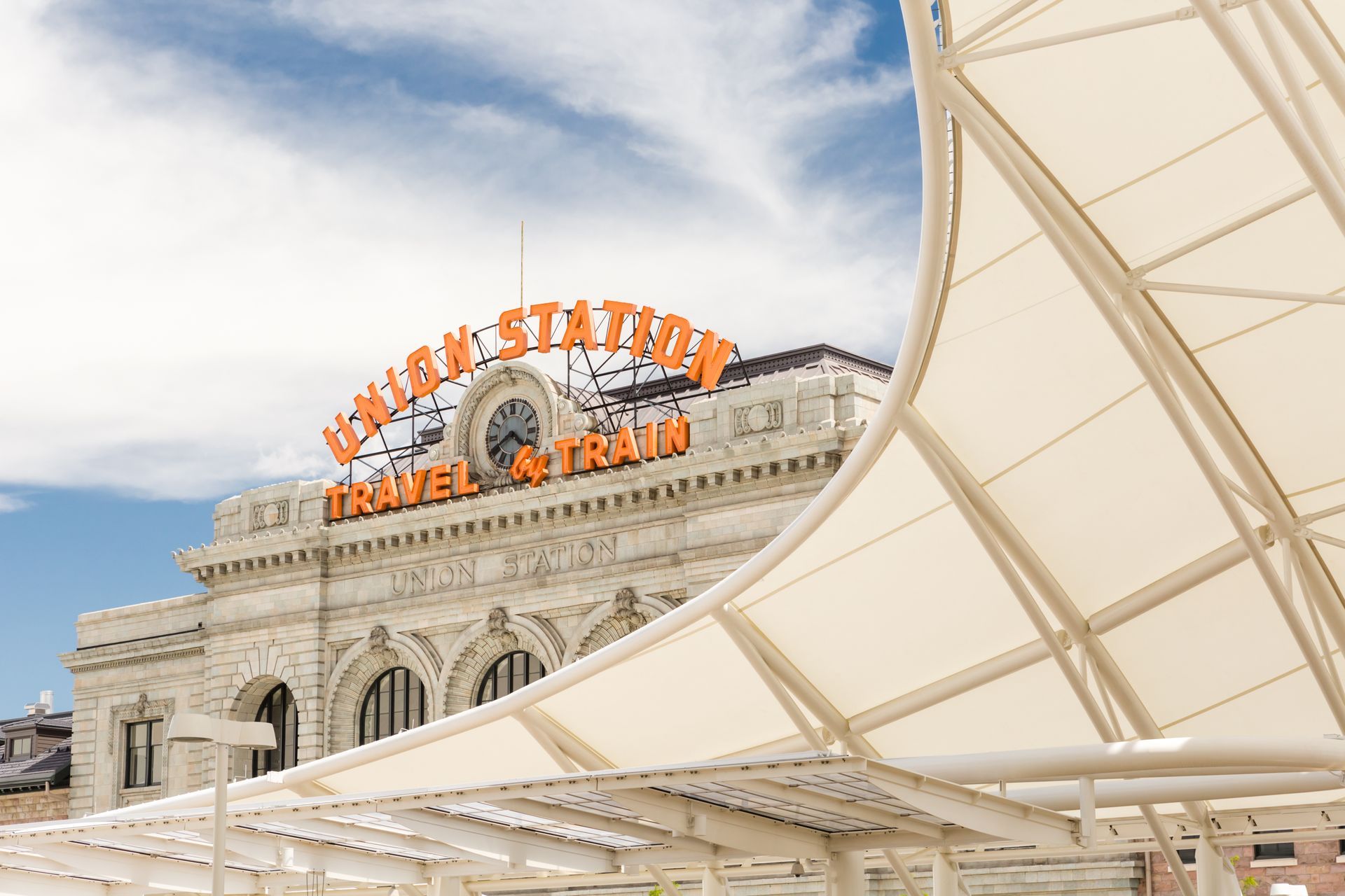 Union Station in Denver, Colorado, with large orange sign. Cream colored awning in foreground, blue sky with clouds.