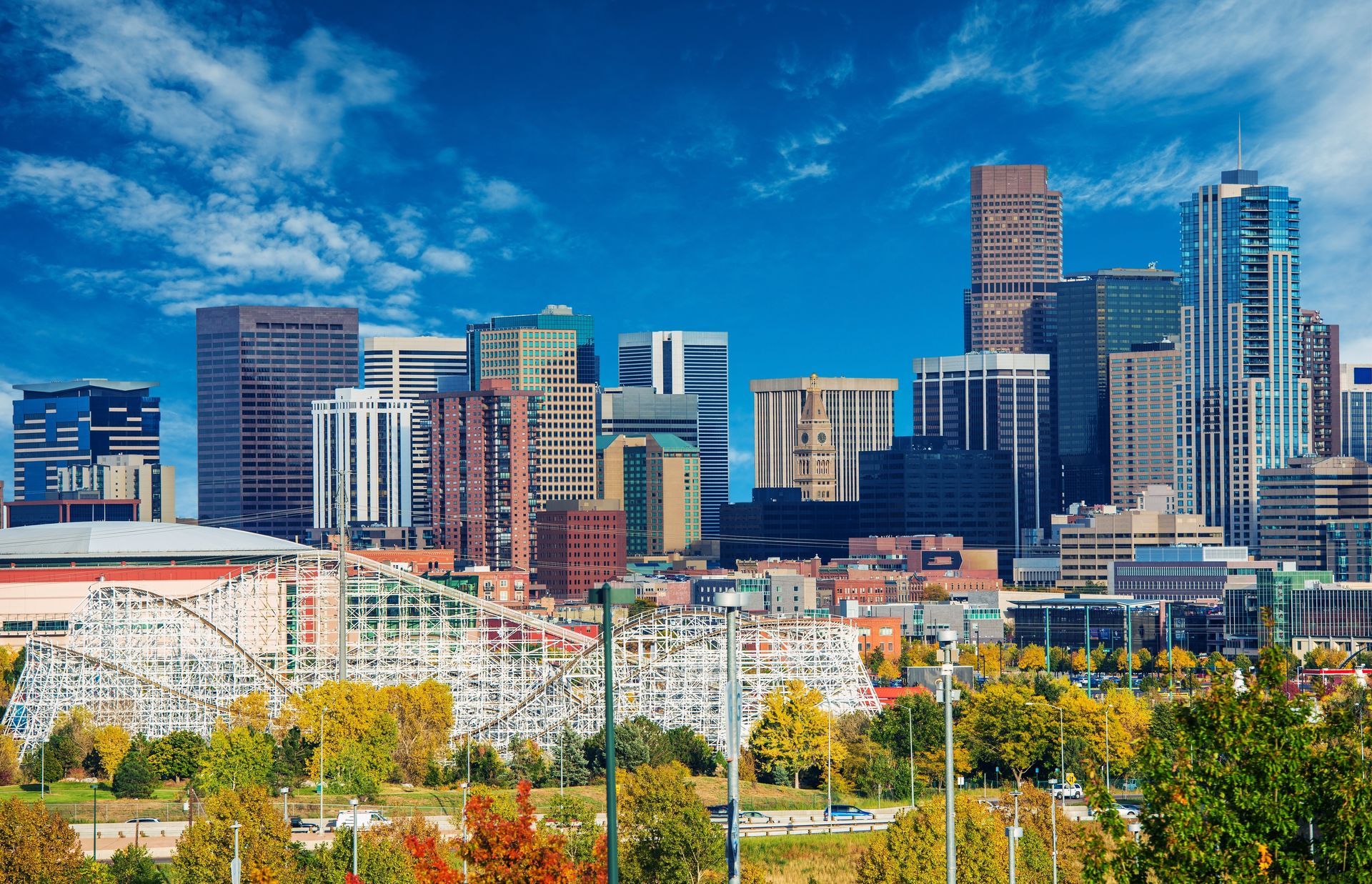 Downtown Denver skyline with various buildings against a blue sky, trees in foreground.