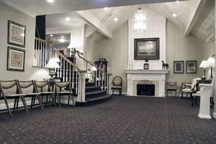 Funeral home interior, grand staircase, fireplace, chairs, carpeted floor, and art.