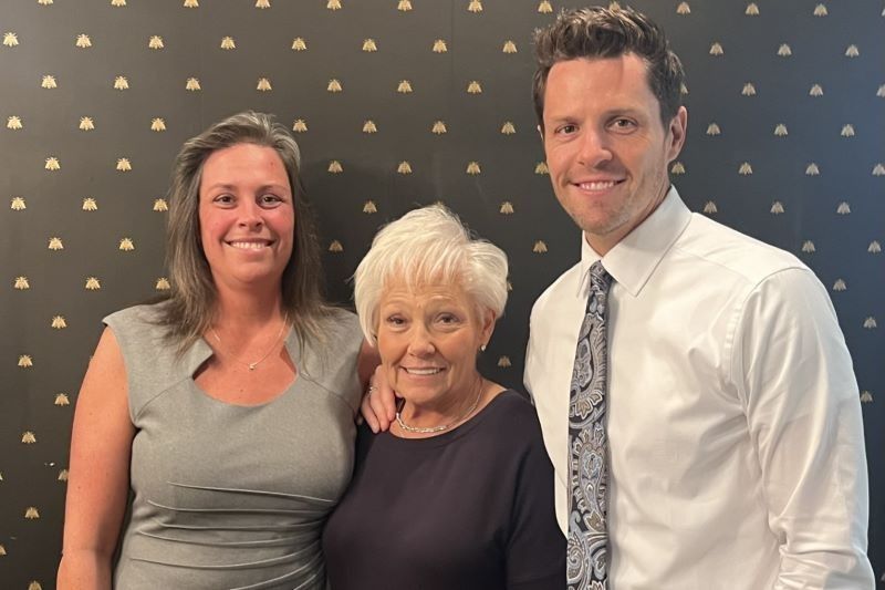 Three people smiling, standing in front of a black patterned wall. Woman in gray, older woman in black, man in white shirt.