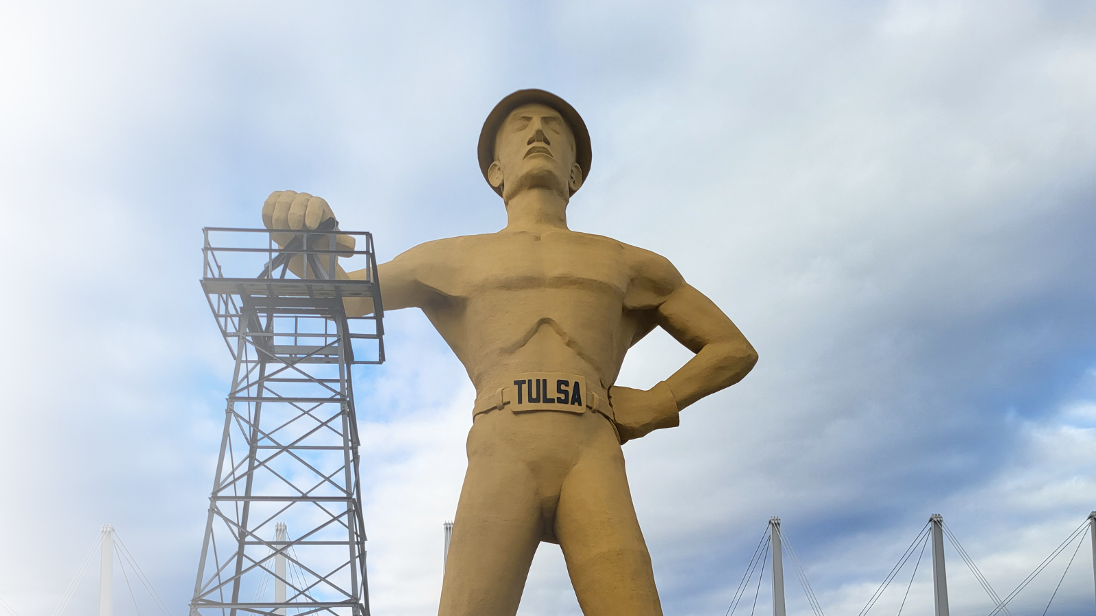 Golden Driller statue in Tulsa, Oklahoma, with a tower behind it and a cloudy sky.