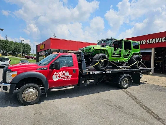 Red tow truck carrying a green off-road vehicle outside an auto service shop