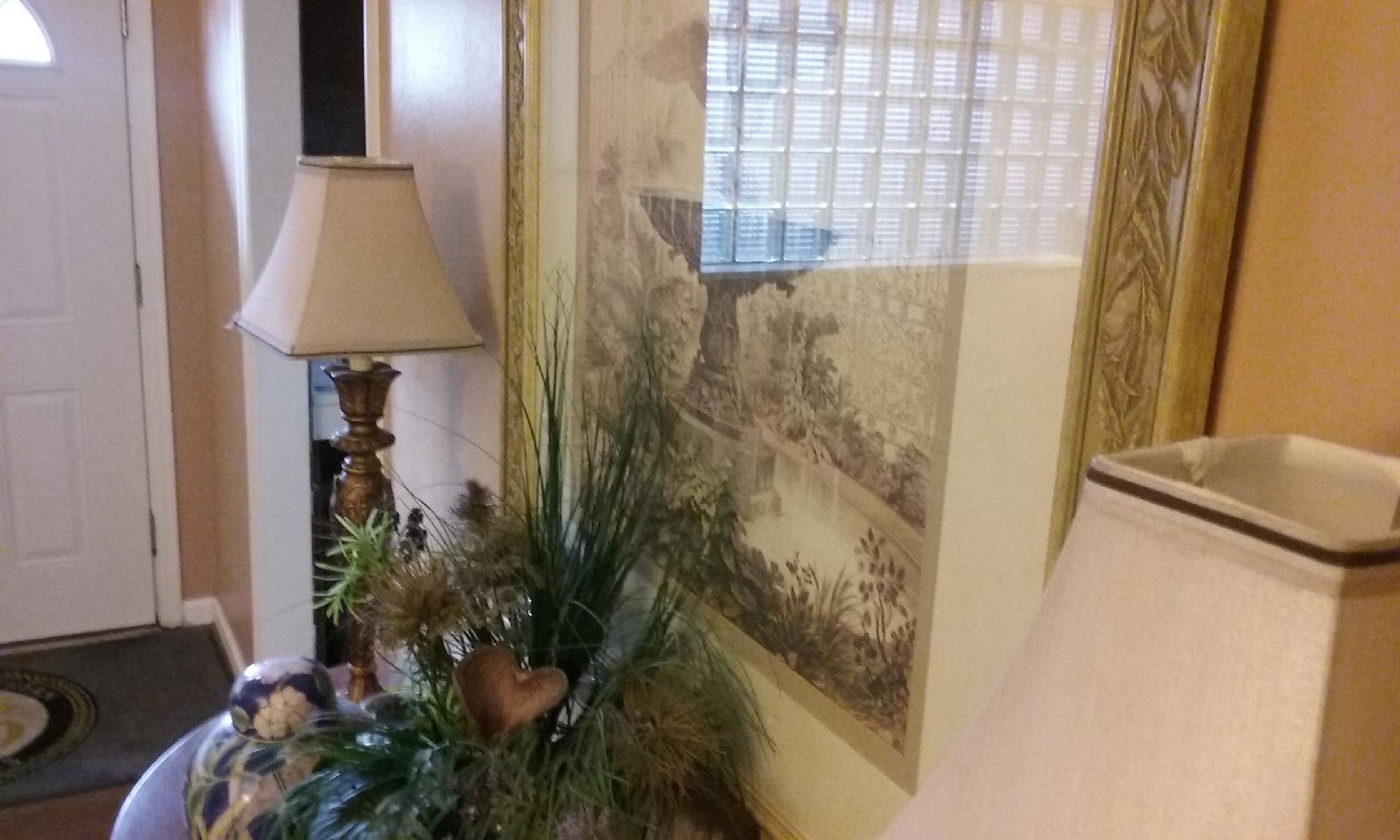 Lamp with beige shade next to artwork with a landscape depiction, in a room with a white door.