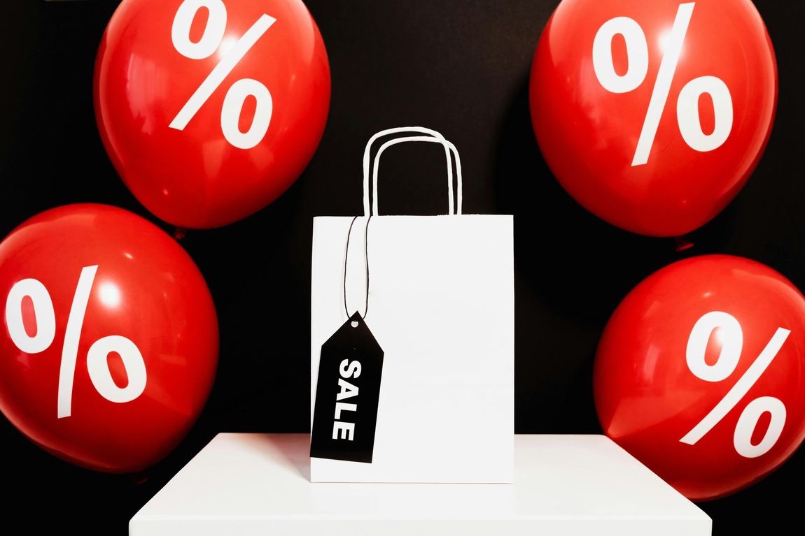 A white shopping bag with a sale tag is surrounded by red percent balloons.