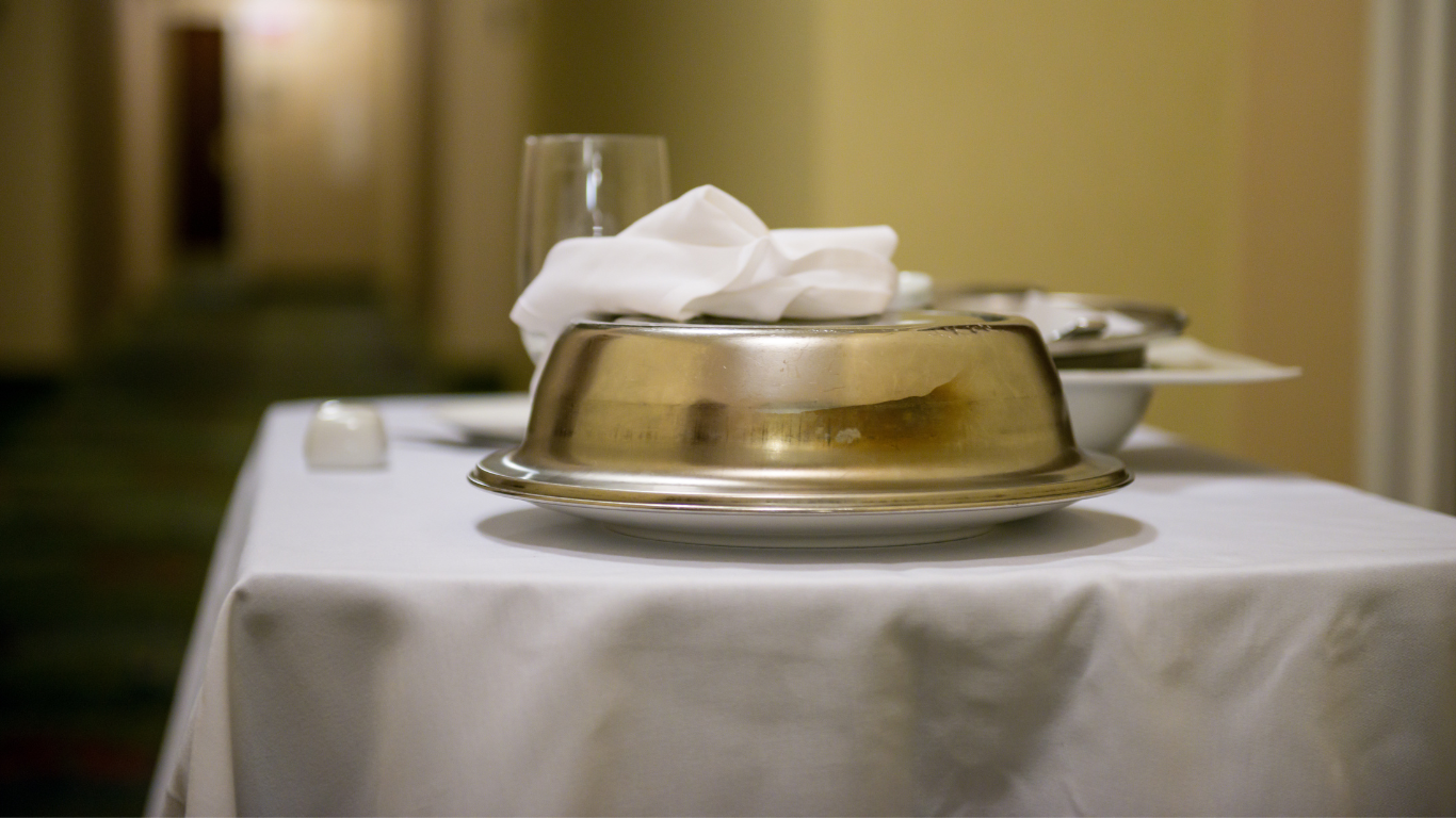 A covered metal serving dish with a white cloth napkin sits on a white-clothed hotel room service cart in a hallway.