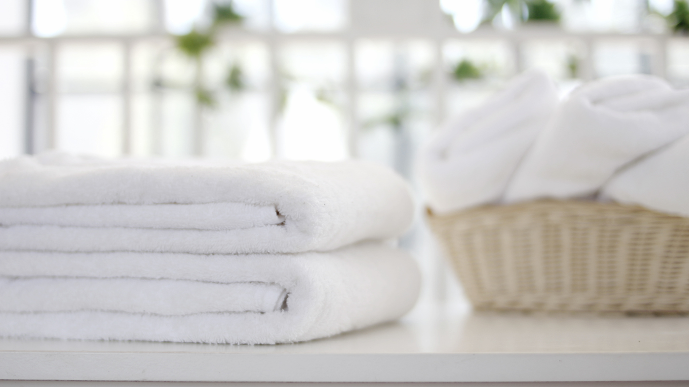 A stack of neatly folded white towels next to a wicker basket filled with more white towels on a light surface.