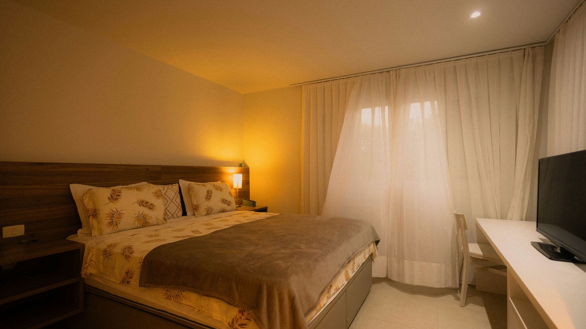 Bedroom with a bed, nightstand, desk, and window with curtains; lit by warm light.