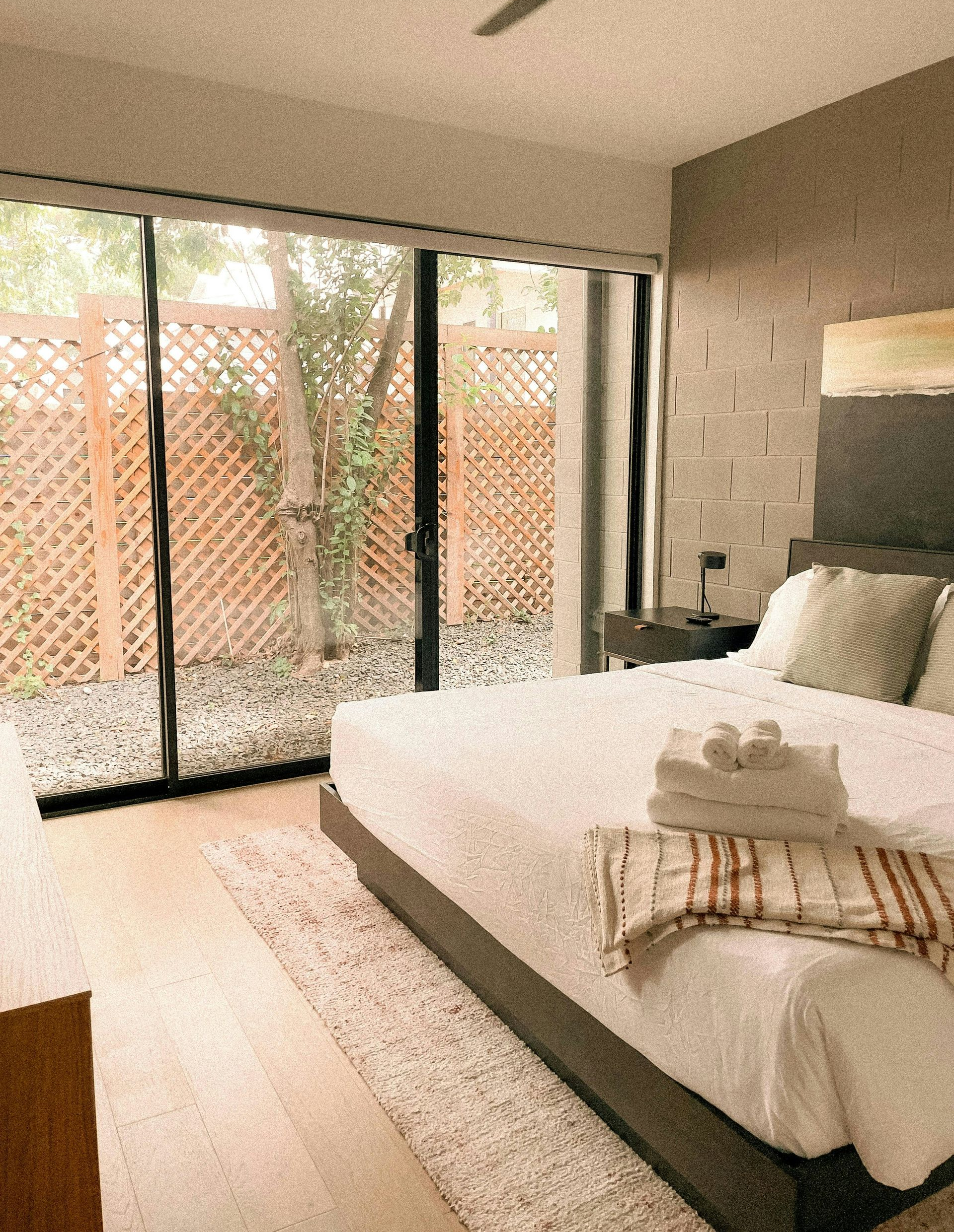 Modern hotel room with a large bed, light wood floors, and a floor-to-ceiling glass door leading to a patio with a tree.
