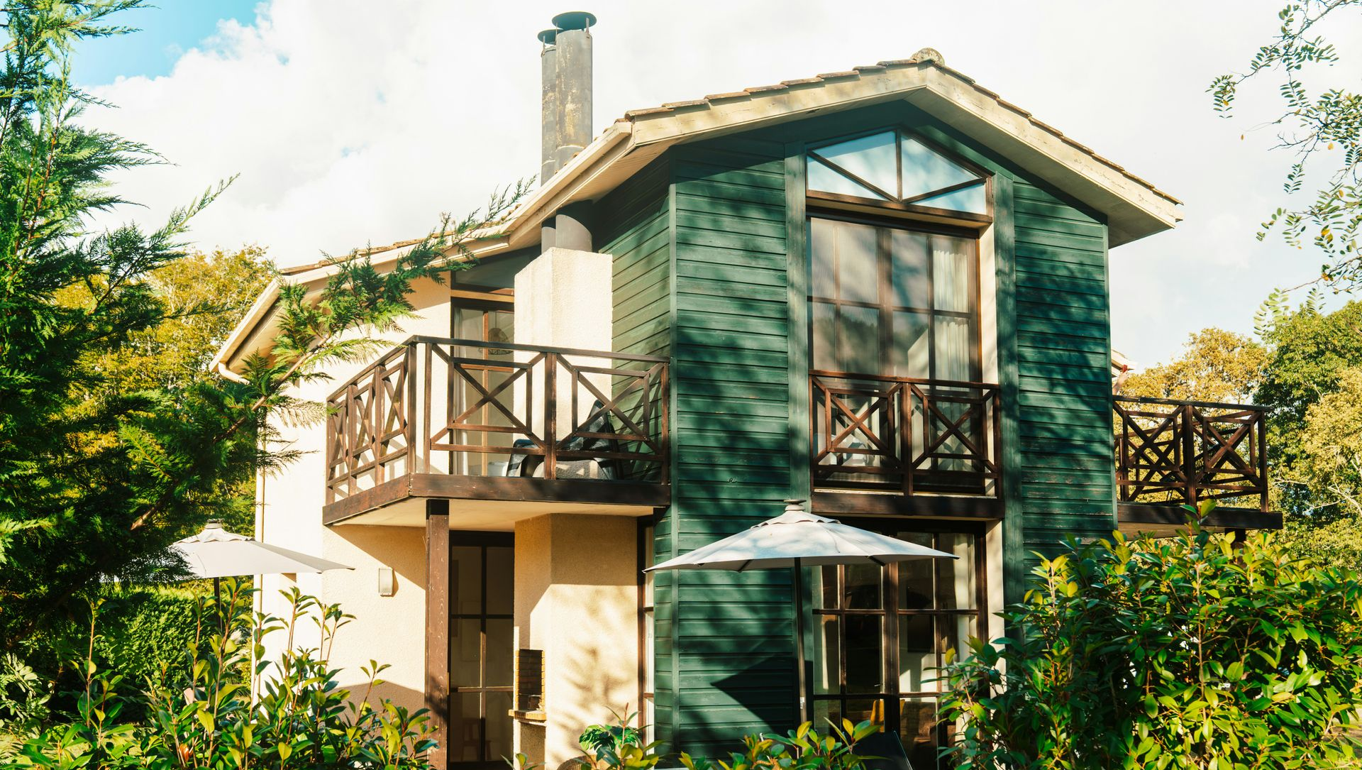 A two-story house with green siding and wooden balconies set amidst lush trees and greenery.
