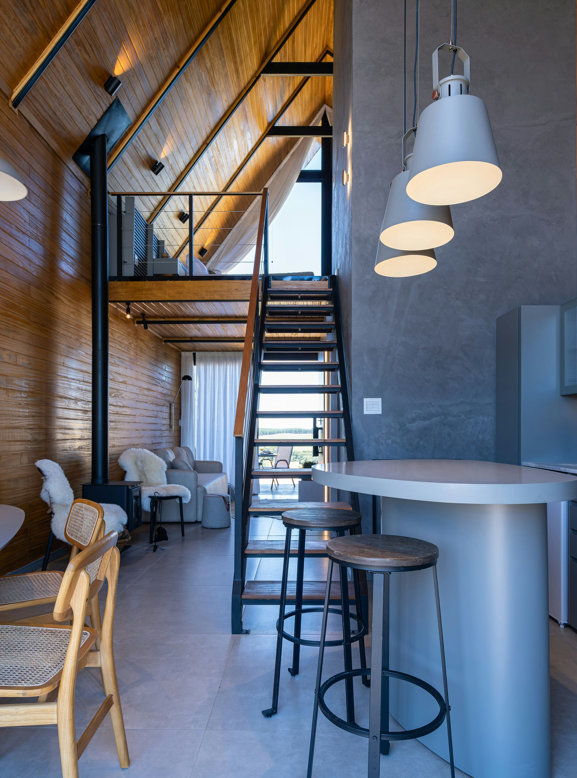A modern, open-concept living space with a wooden A-frame ceiling, a loft area, a metal staircase, and a breakfast bar.