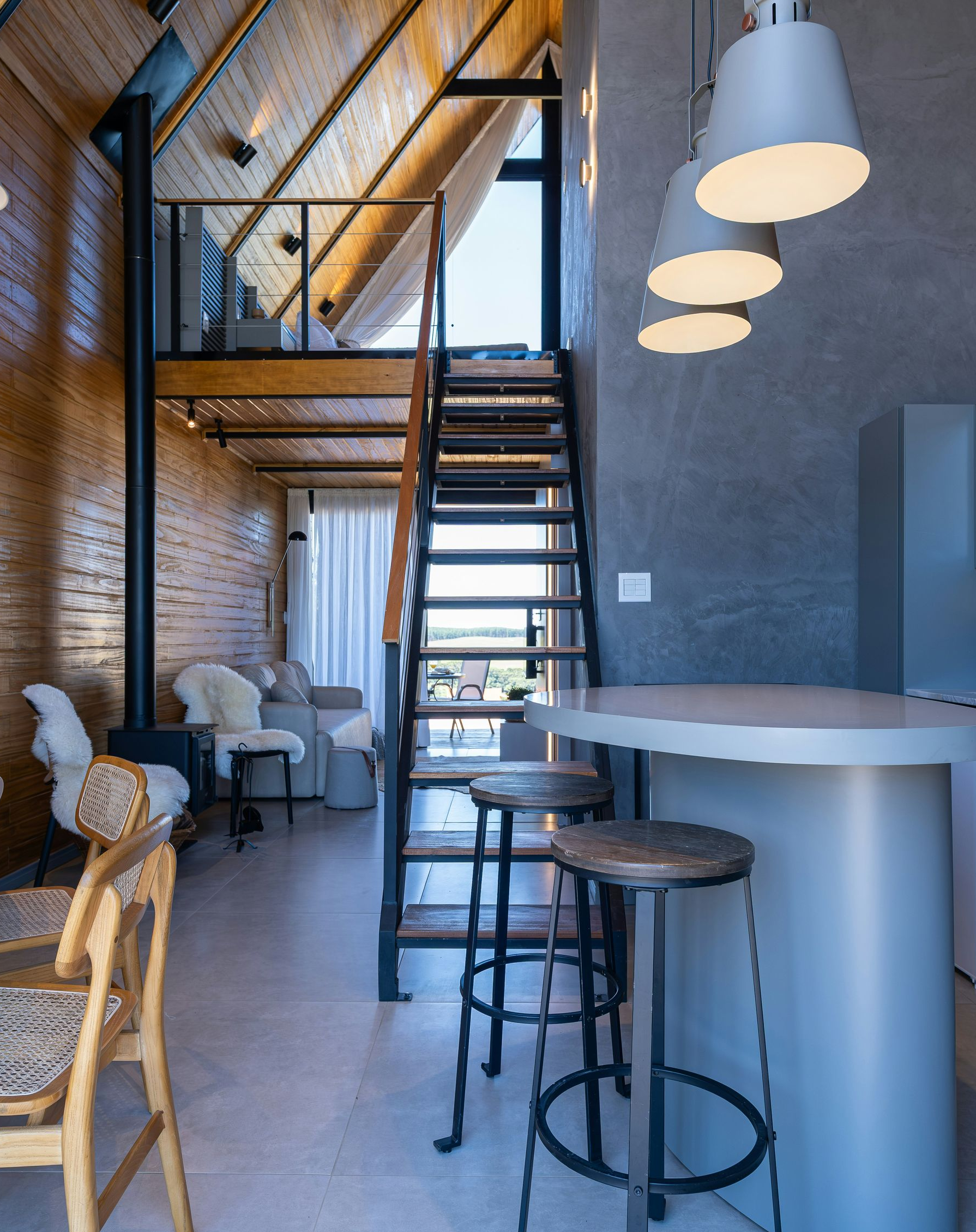 A modern, open-concept living space with a wooden A-frame ceiling, a loft area, a metal staircase, and a breakfast bar.