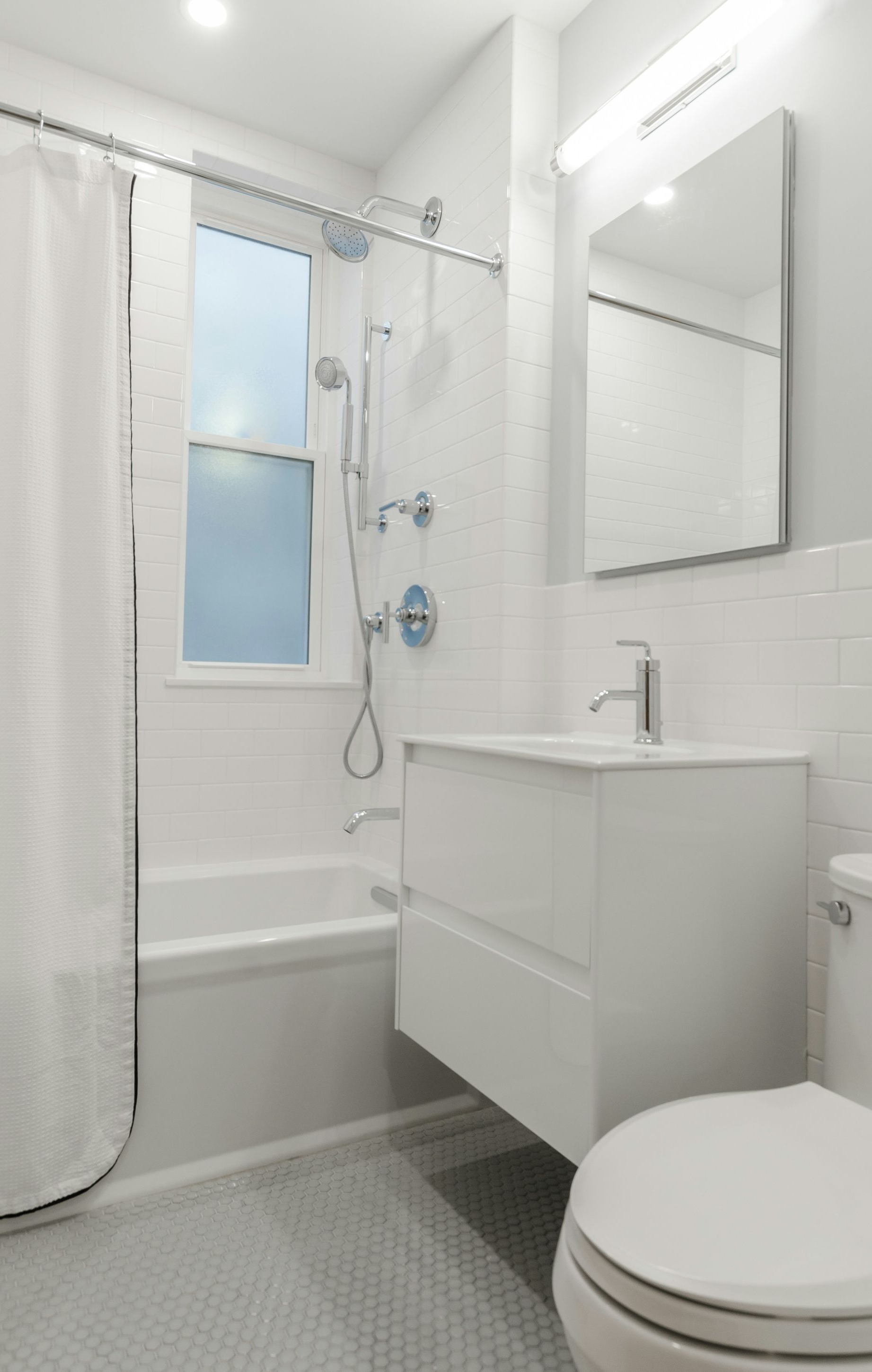 White bathroom with shower, vanity, toilet, and window; gray tiled floor.