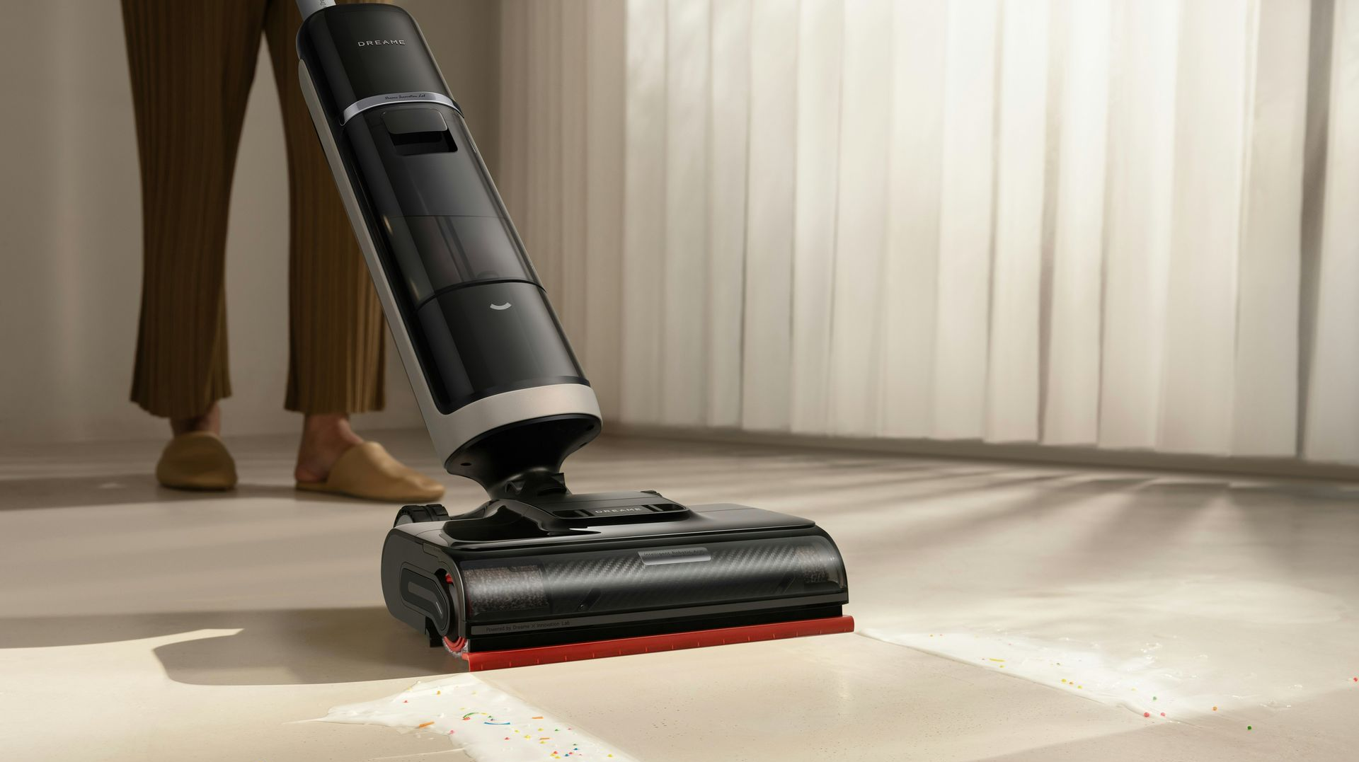 Person using a black and silver floor cleaner on a light-colored floor, near a white curtain.