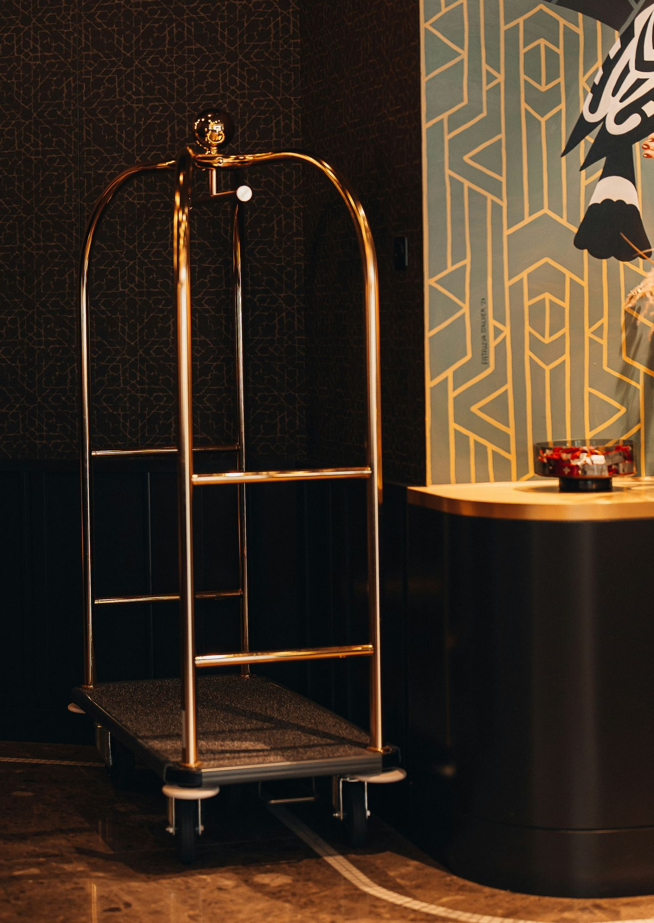 A gold metal luggage cart stands next to a wall with black textured wallpaper and geometric, Art Deco-style patterns.