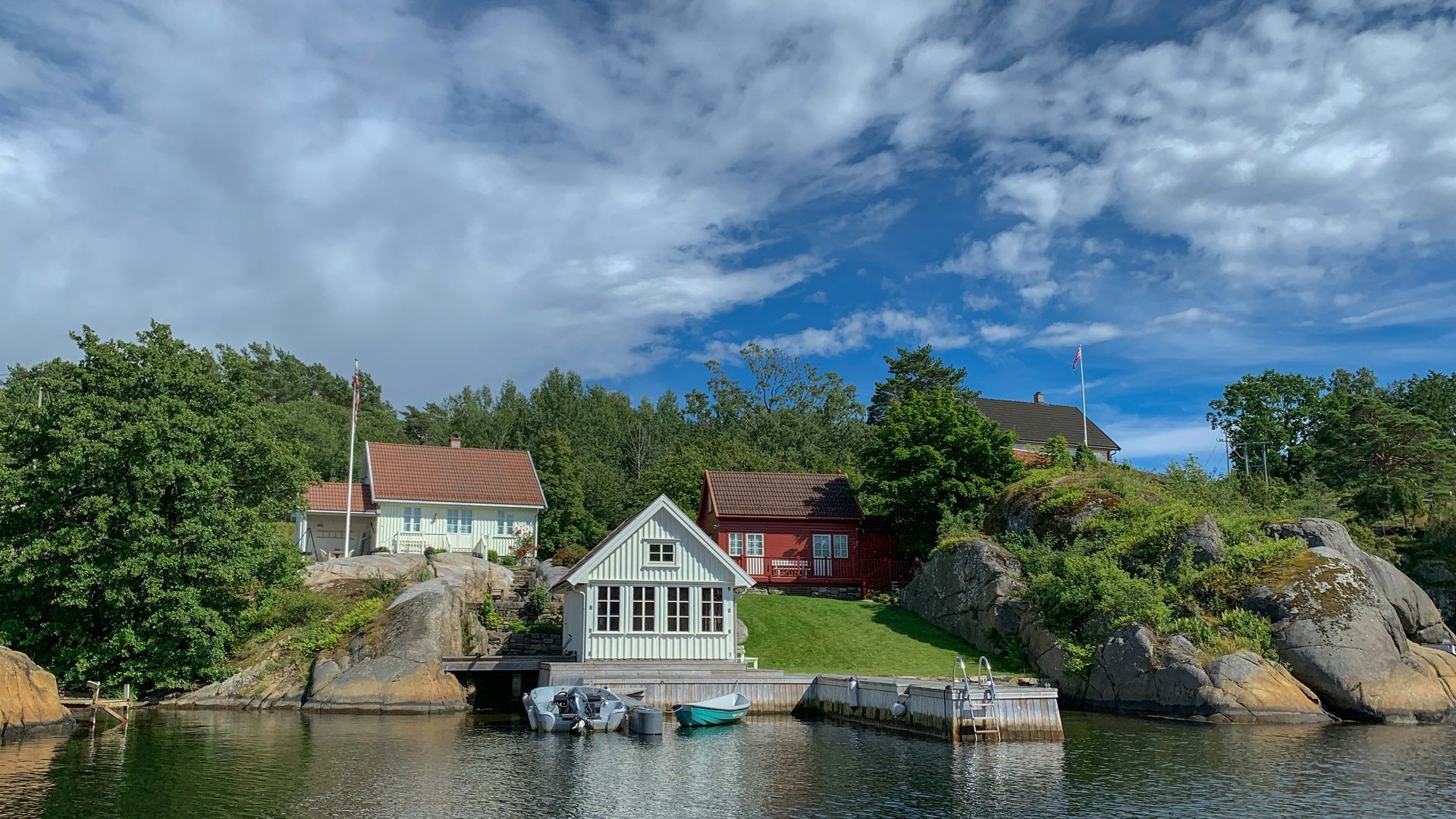 Houses on rocky shoreline, green trees, cloudy sky, boats at dock.
