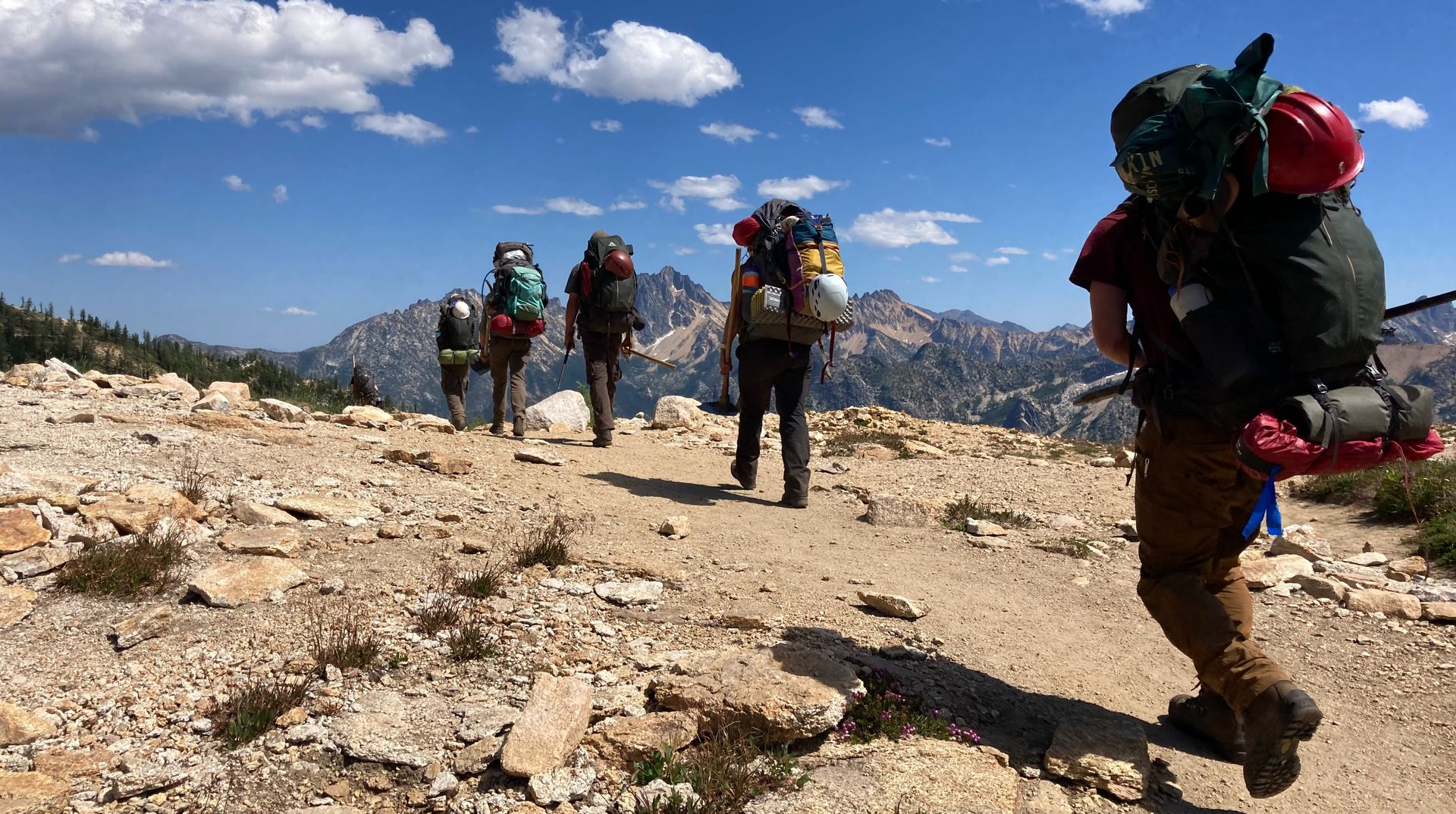 Hikers with backpacks crossing a rocky mountain trail under a bright blue sky with clouds
