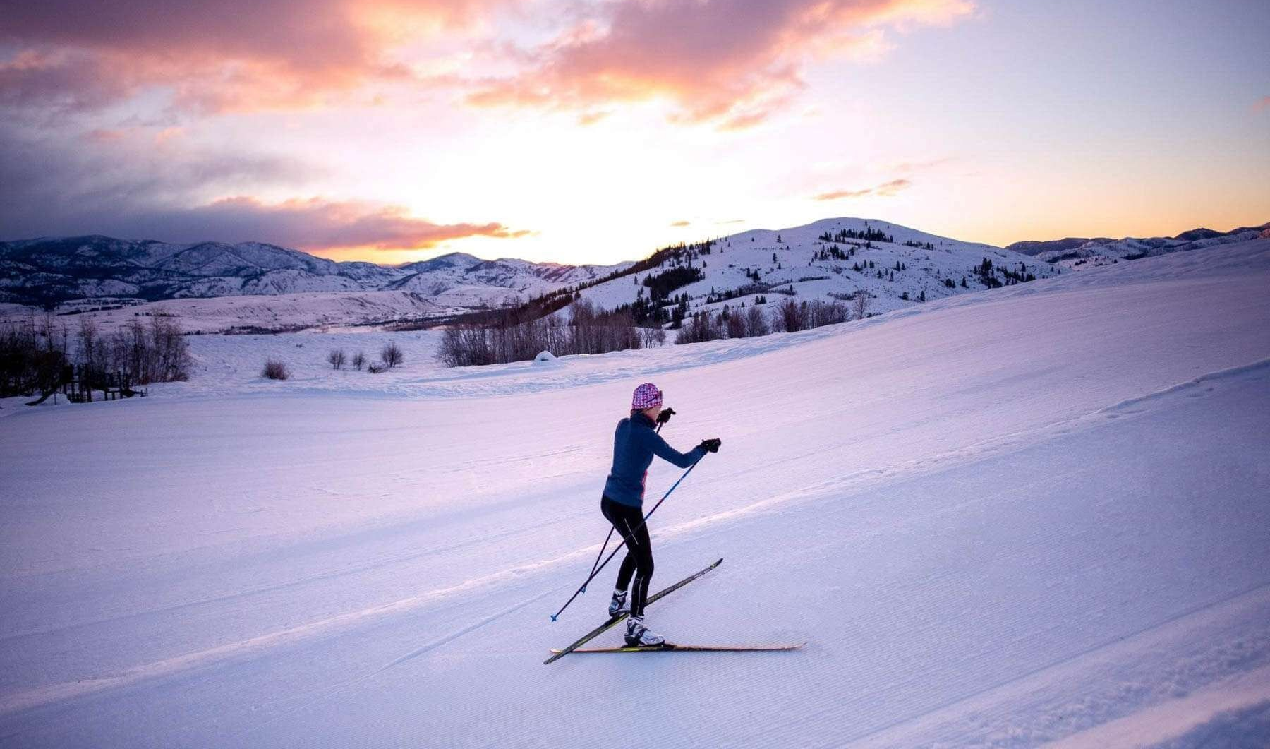 Skier gliding across a snowy slope at sunset with mountains in the background