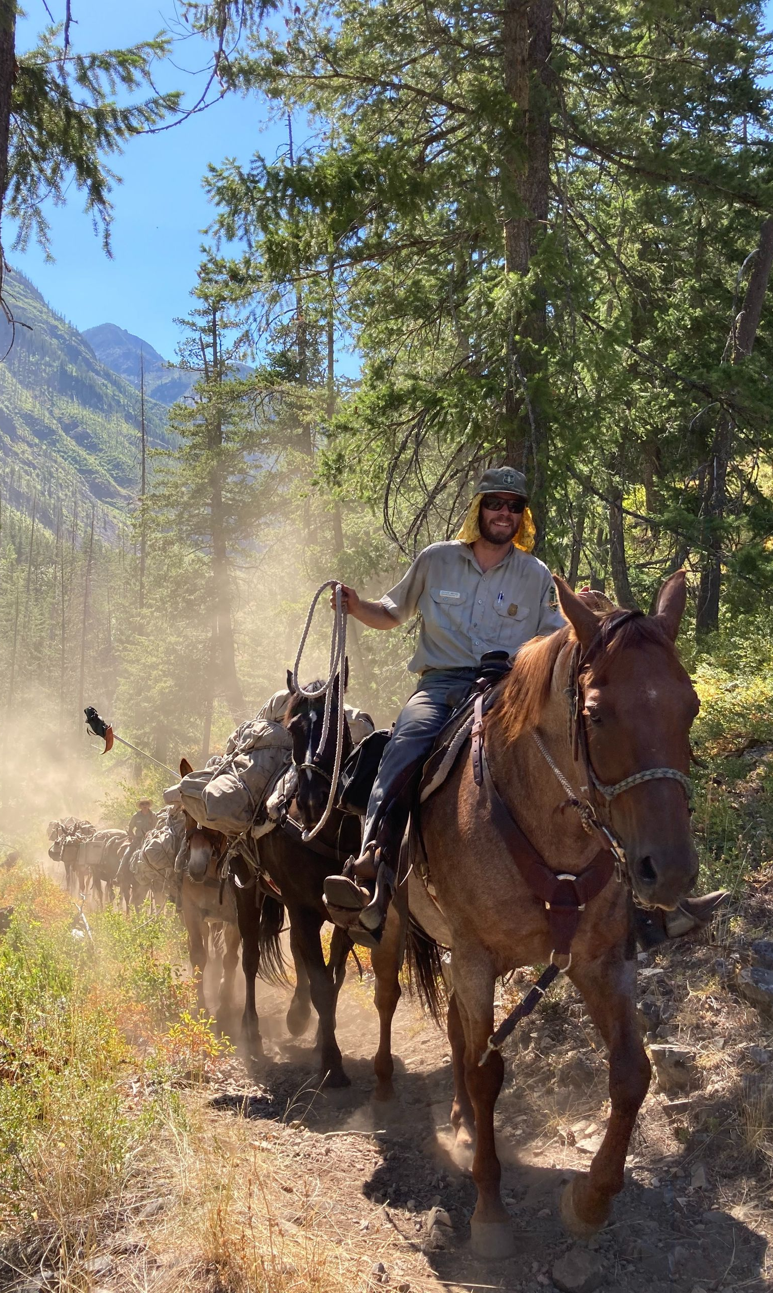 Person riding a horse through a dusty forest trail with mountains in the background