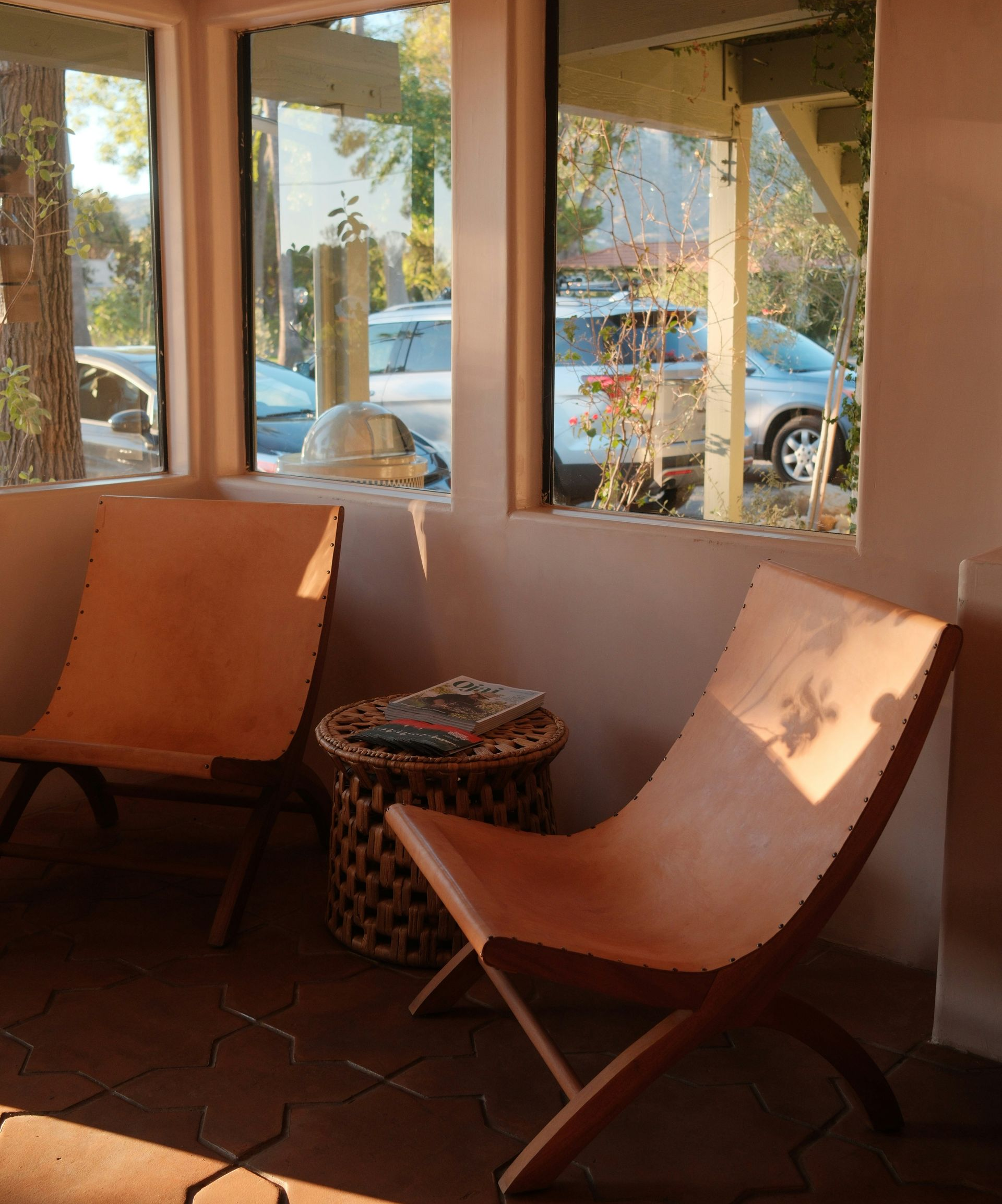Two wooden chairs with leather seats in a sunlit room, next to a wicker table, looking out at vehicles.