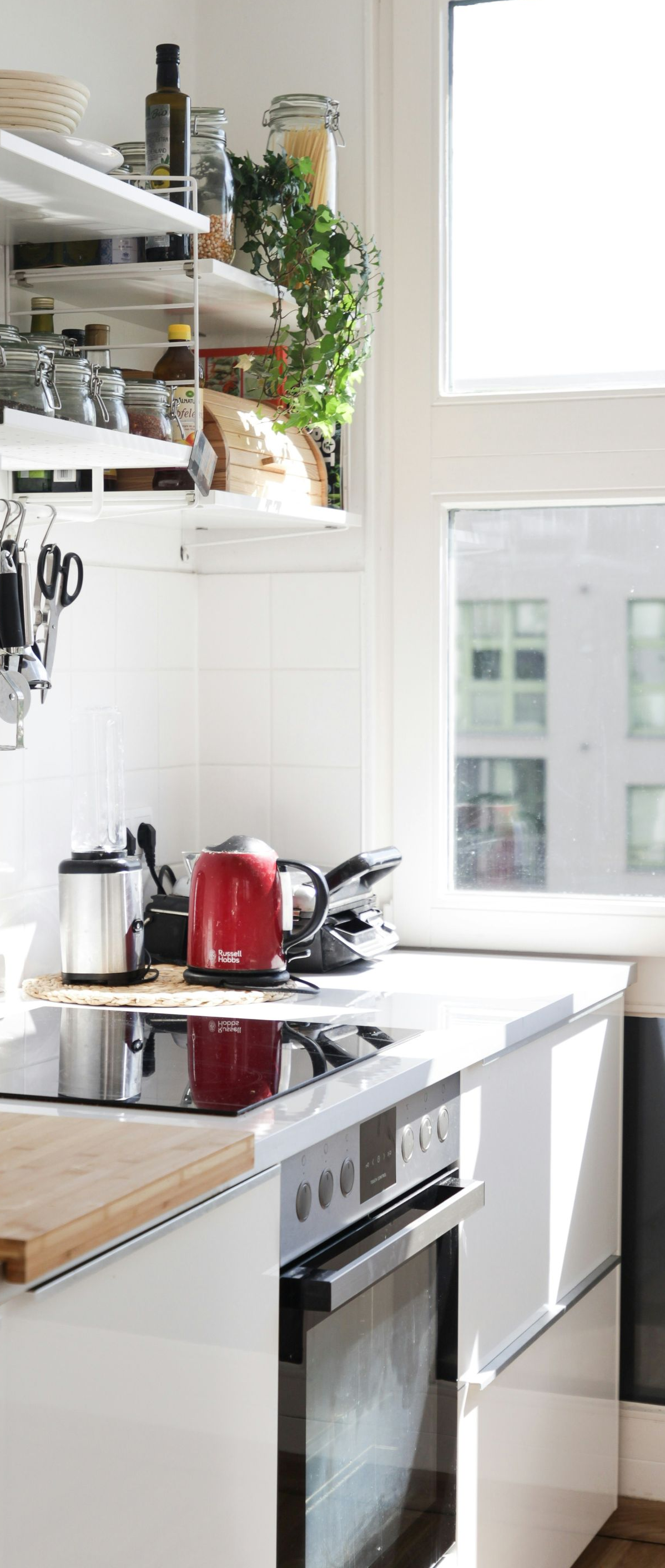 Bright white kitchen with open shelves, wooden countertop, red kettle, and sunlight by a window.