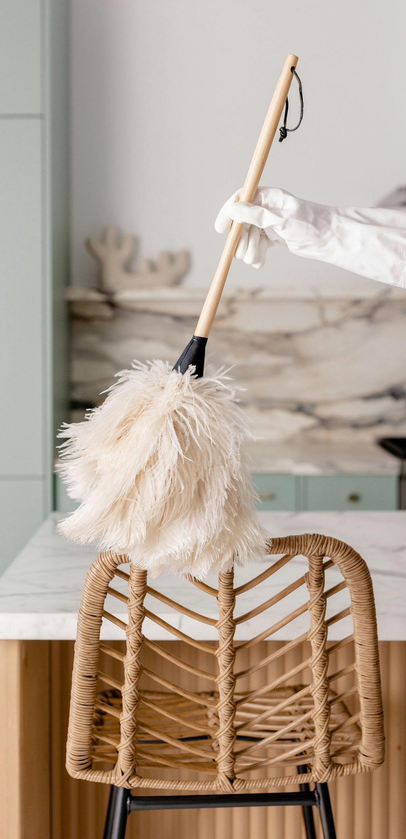 Person in glove dusts a woven chair with a feather duster in a kitchen with marble countertop.