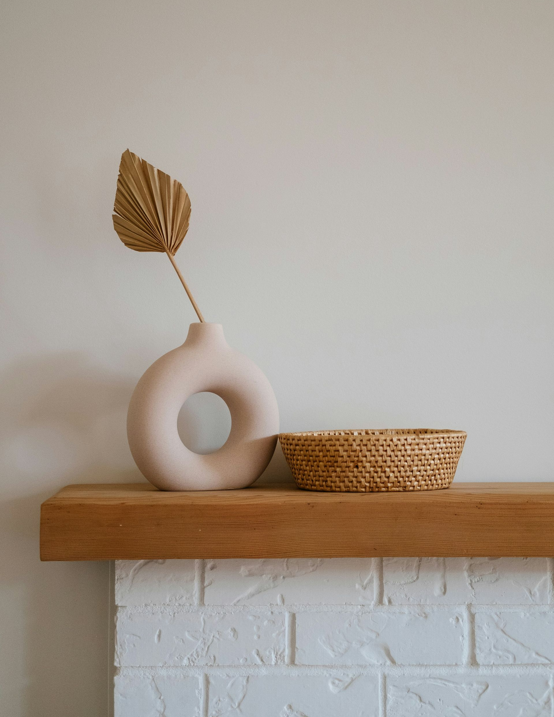 A neutral-colored donut vase holding a single gold palm leaf next to a small woven basket on a wooden mantelpiece.