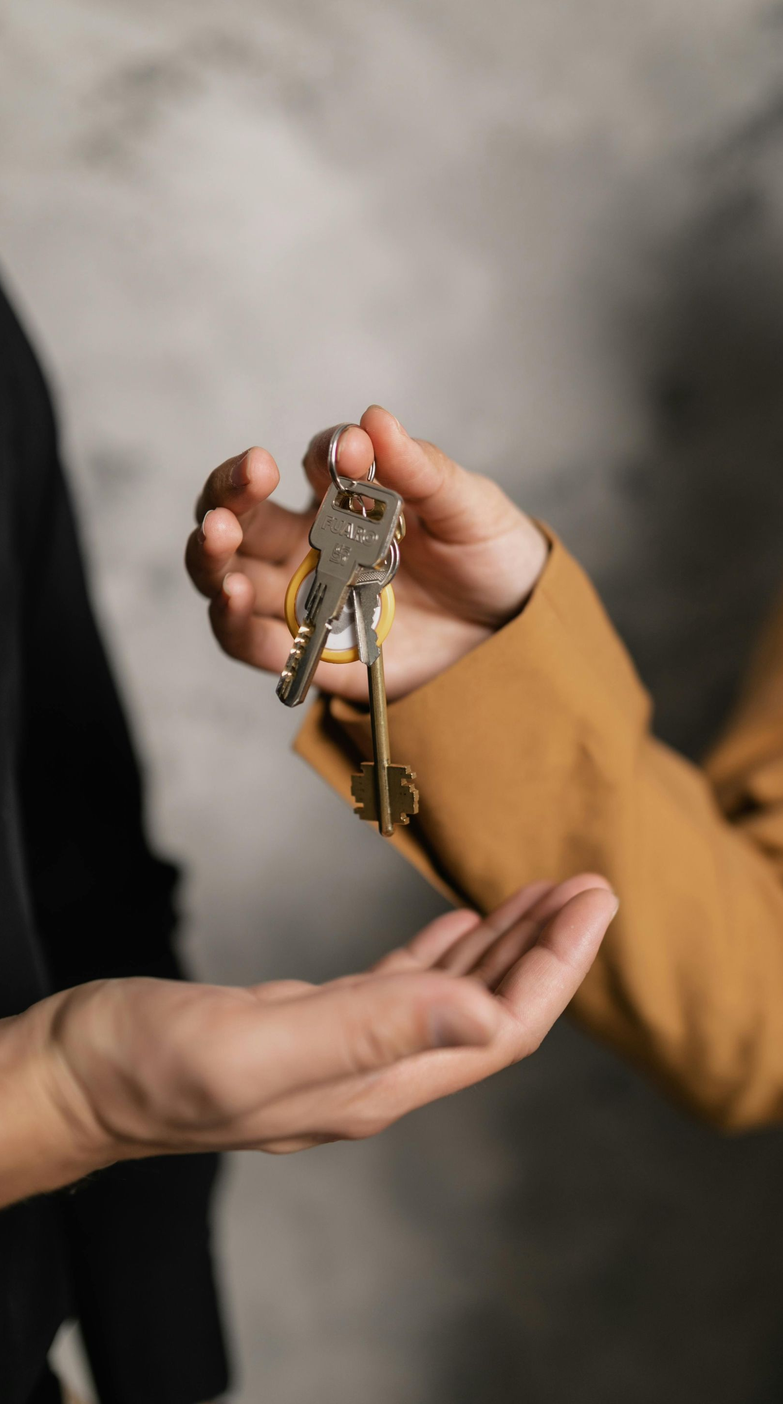 Person's hands exchanging keys. One wearing a tan jacket, the other wearing a black top. Against a gray background.