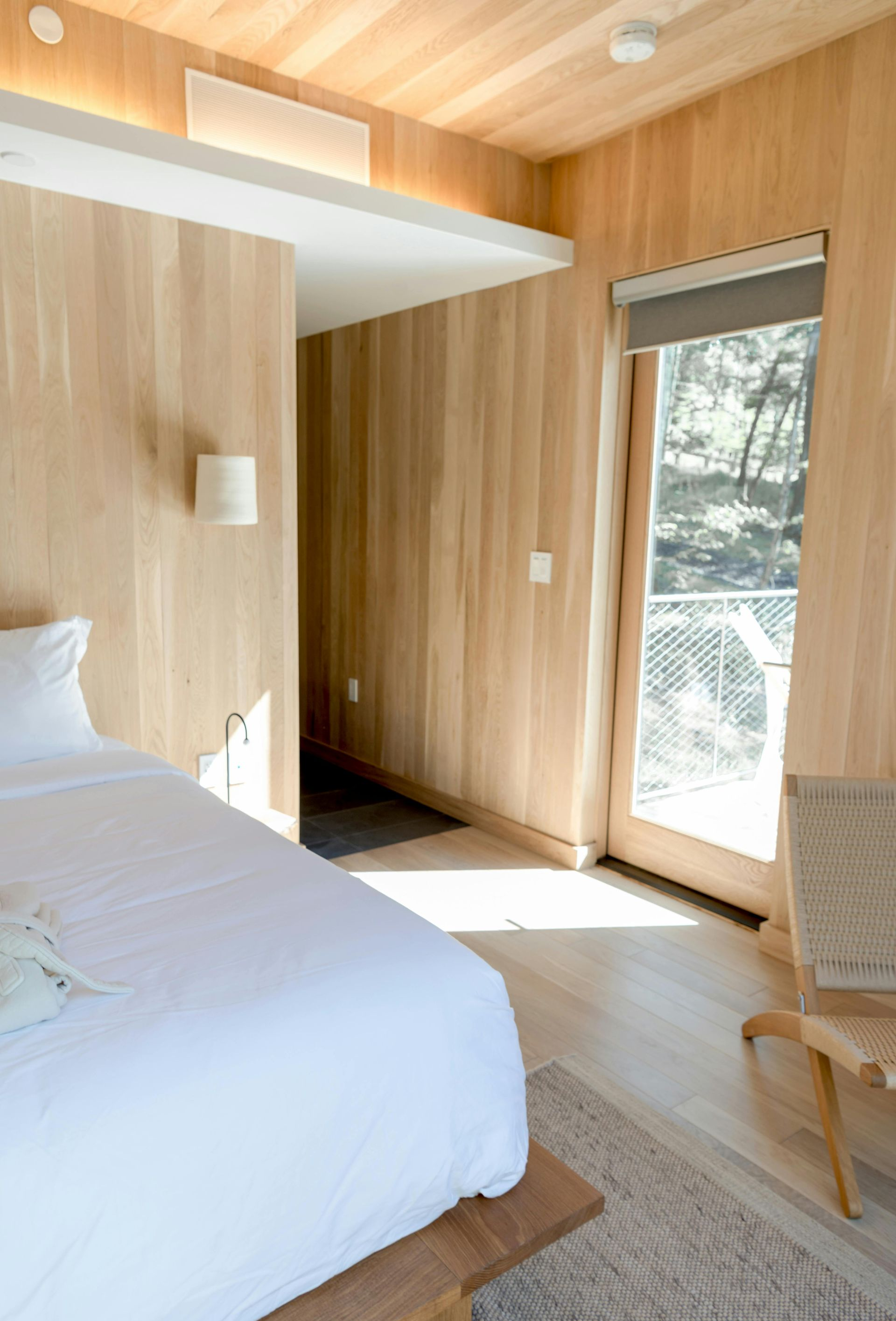 Bedroom with light wood paneling, white bedding, and a doorway to a balcony. Natural light streams in.