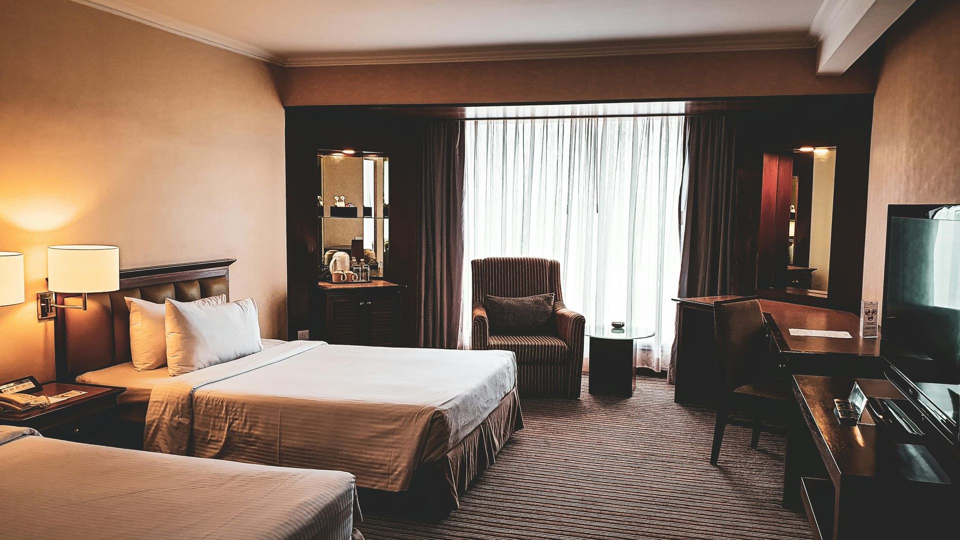 Hotel room with two beds, a chair, and a desk, all in warm tones.