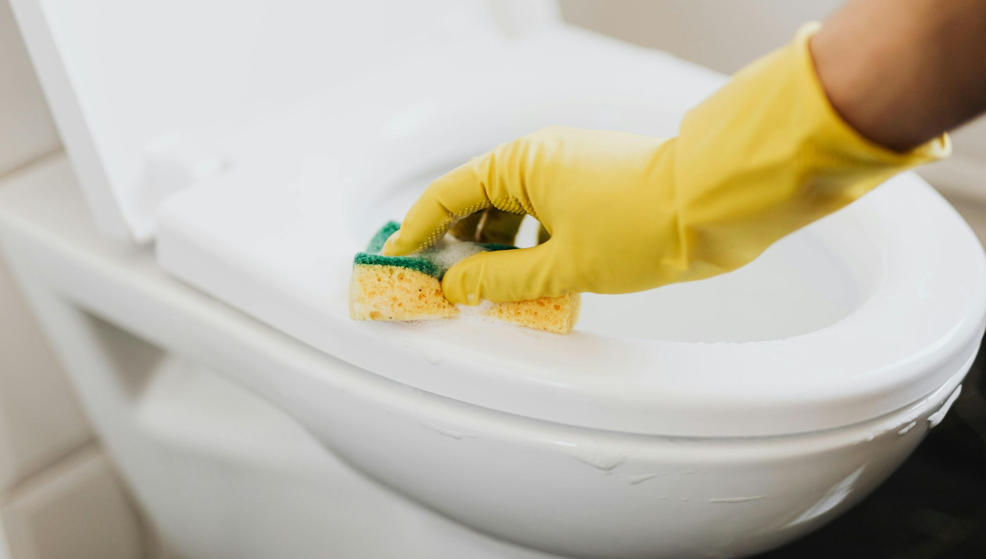 A hand in a yellow rubber glove uses a sponge to scrub the rim of a white toilet.