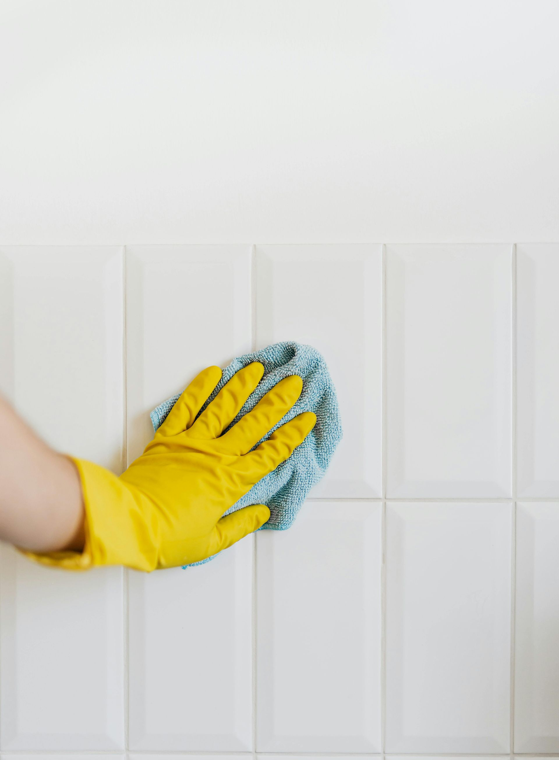 A hand wearing a yellow rubber glove uses a patterned cleaning cloth to wipe down white subway tiles on a wall.