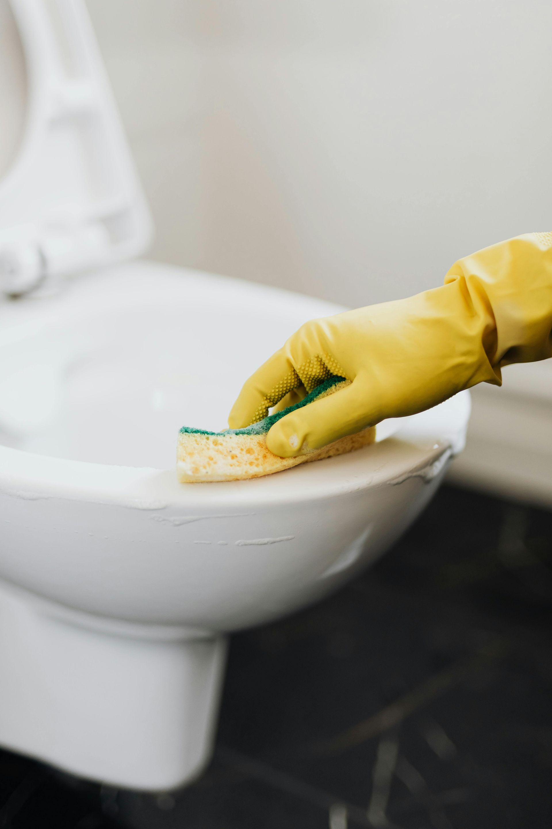 A hand in a yellow rubber glove uses a yellow and green sponge to clean a white toilet.