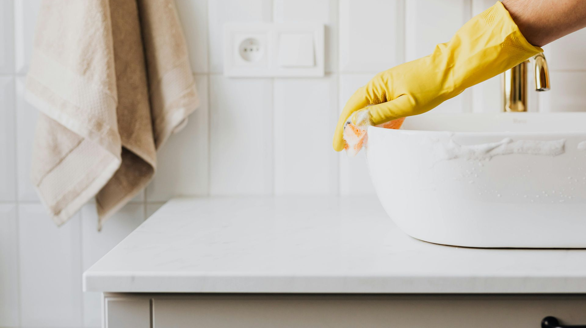 A person wearing yellow rubber gloves wipes a white bathroom sink with a sponge against a tiled wall.