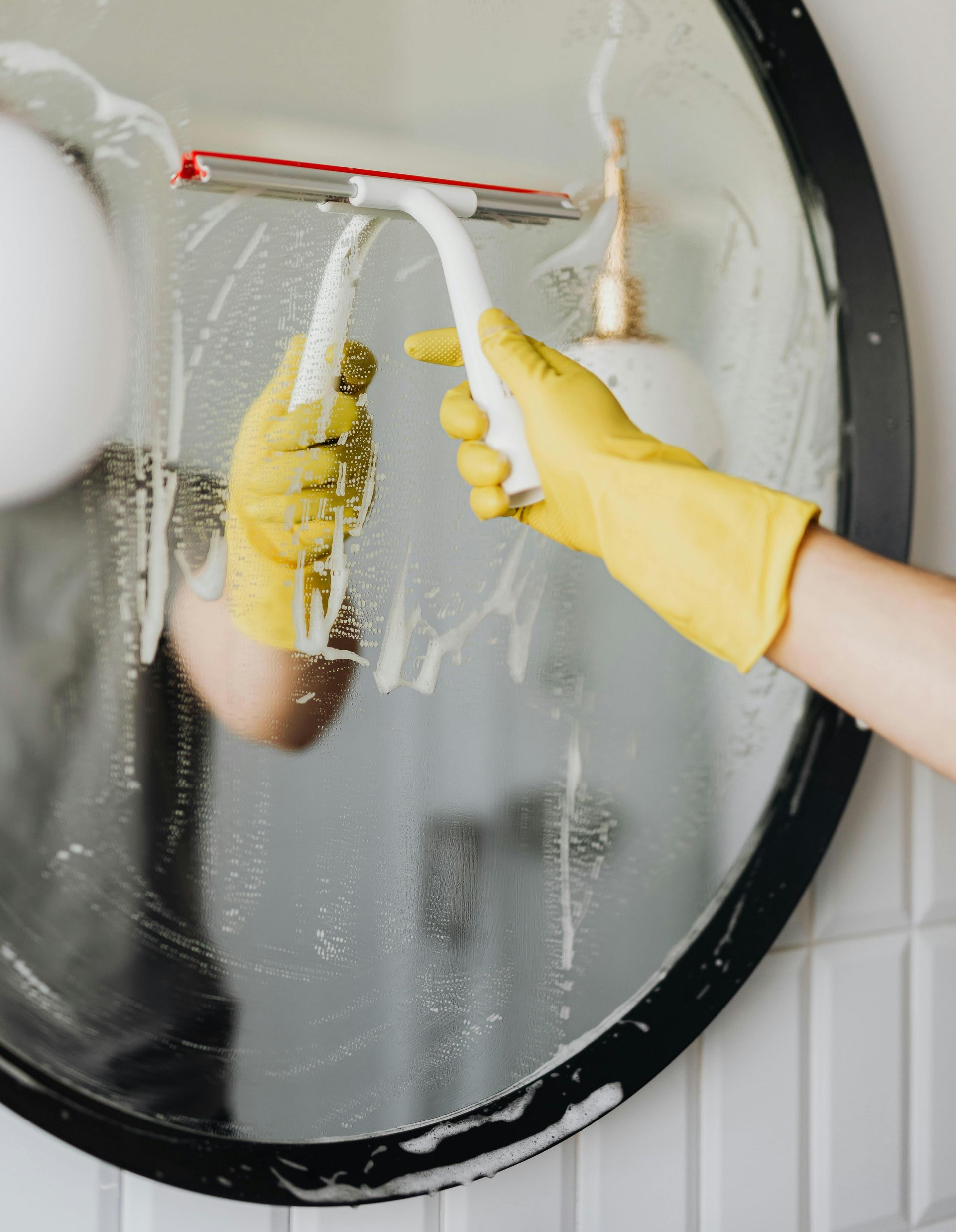 A person wearing yellow rubber gloves uses a squeegee to clean a round, black-framed bathroom mirror.