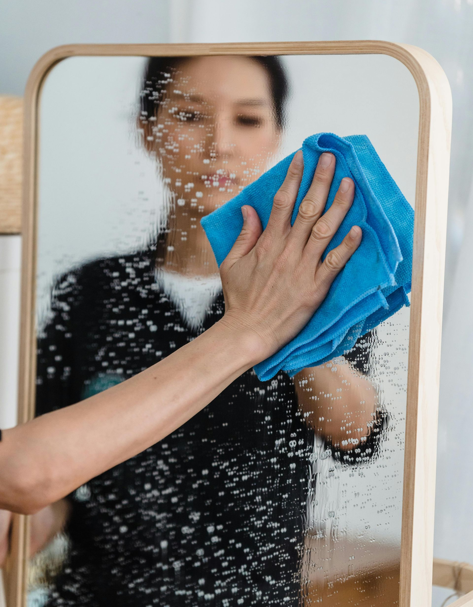 A person wipes a glass mirror with a blue cloth, clearing away small water droplets in a bright, indoor setting.