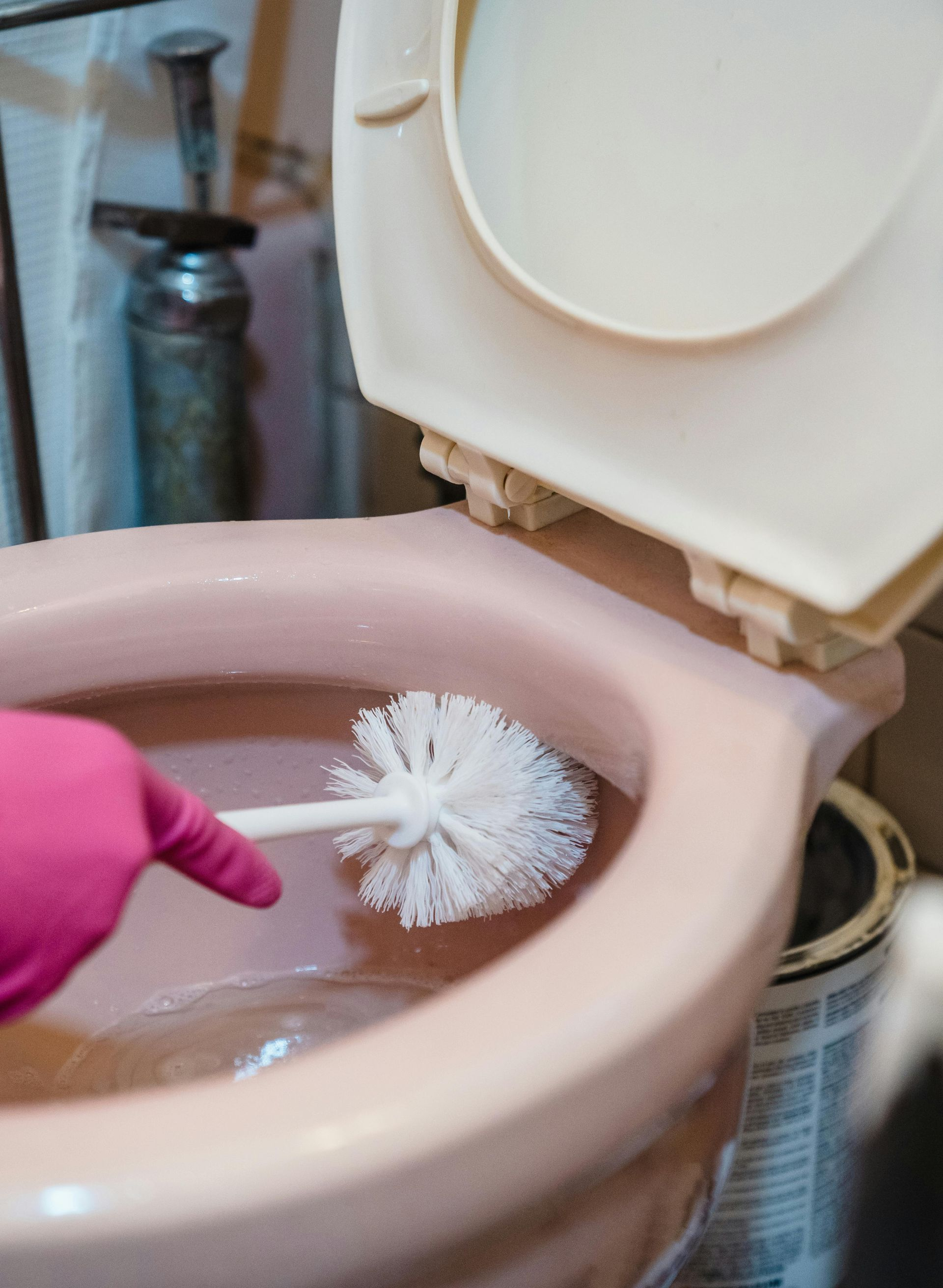 A hand in a pink glove scrubs the inside of a light pink toilet bowl with a white plastic brush.
