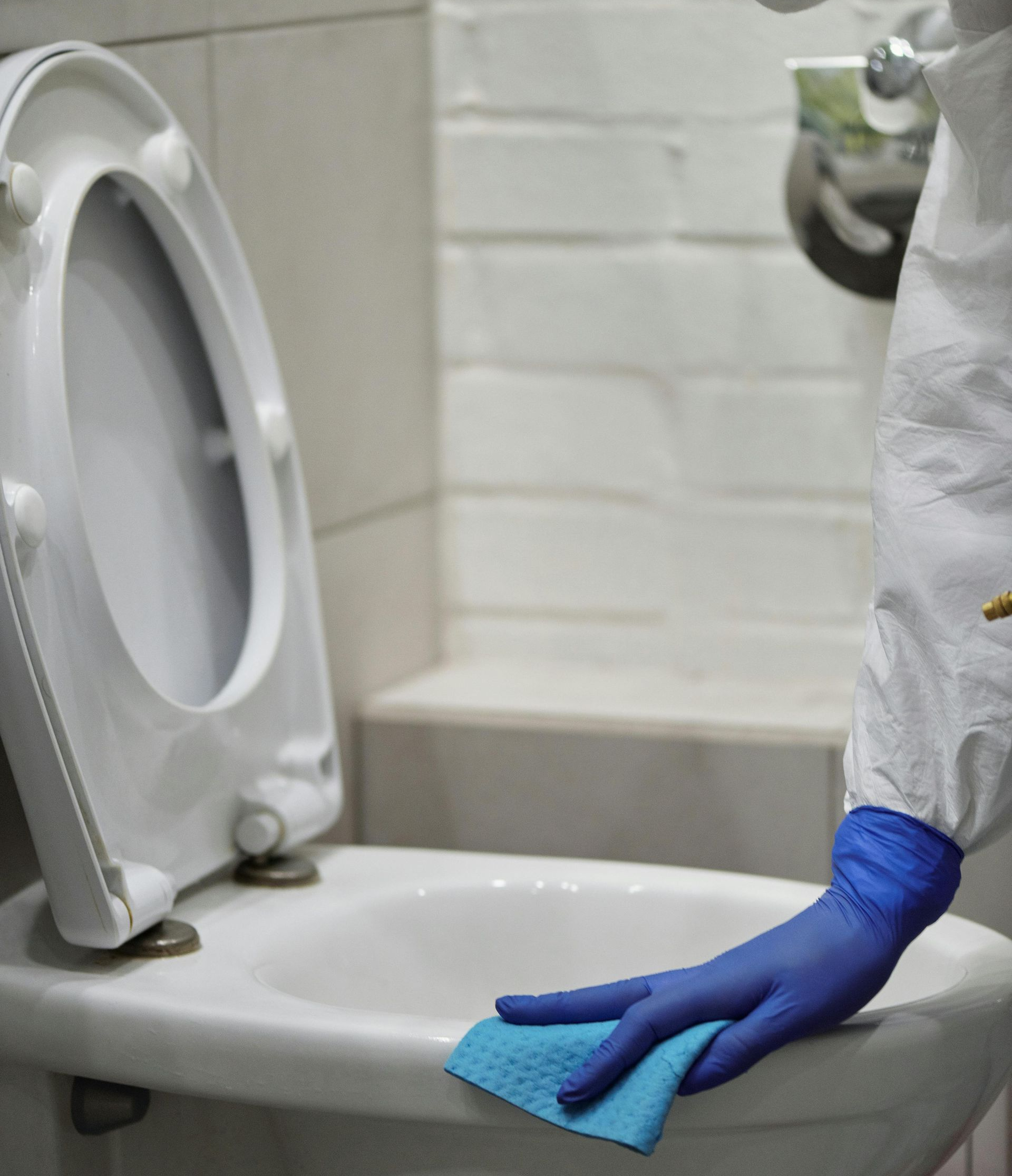 Person in protective suit cleaning a toilet with a blue cloth.