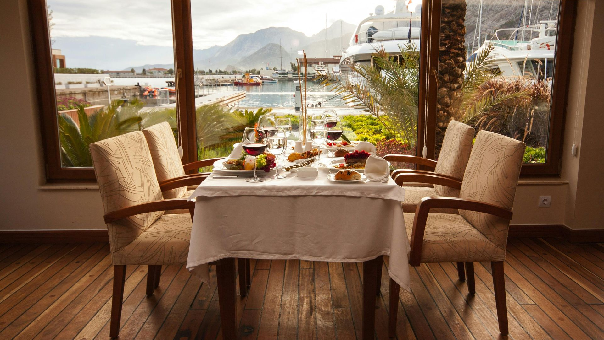 Dining table set for a meal, overlooking a harbor with boats and mountains.