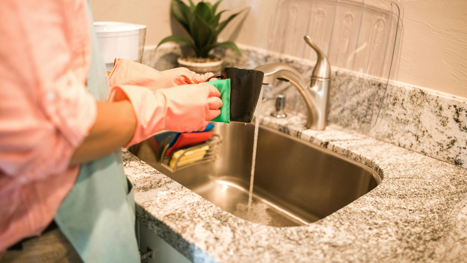 A person in pink sleeves and a blue apron rinses a black object with a green sponge under a kitchen faucet.