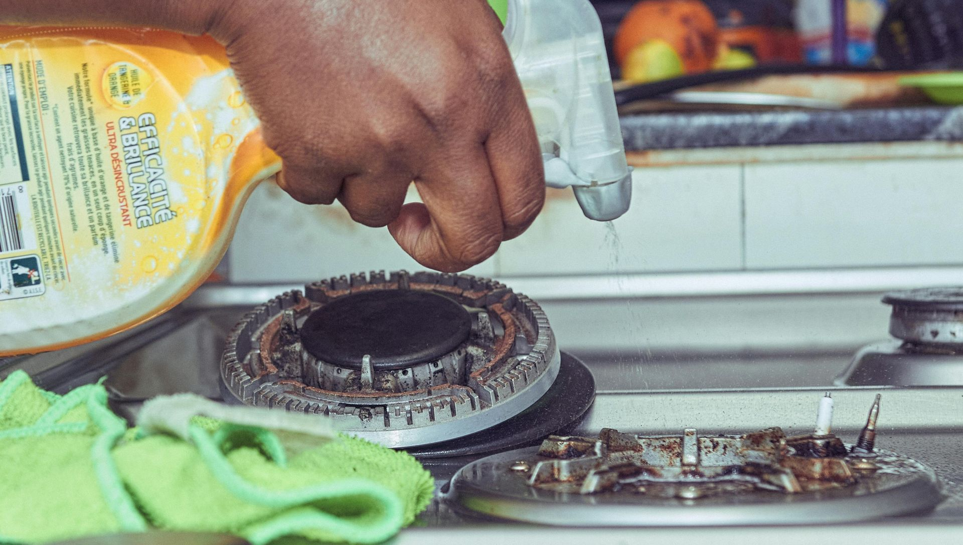 A person sprays a yellow cleaning product onto a gas stovetop, with a green microfiber cloth resting nearby.