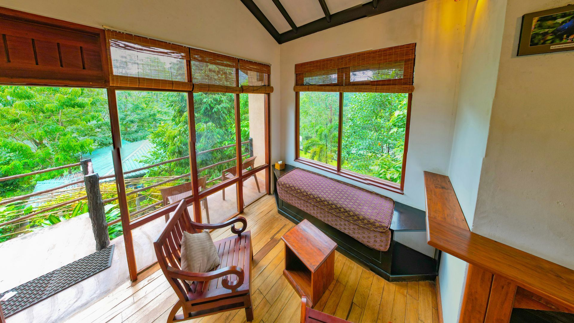 A sunlit lounge with wooden floors, a wooden chair, a built-in bench, and large windows overlooking a lush green landscape.