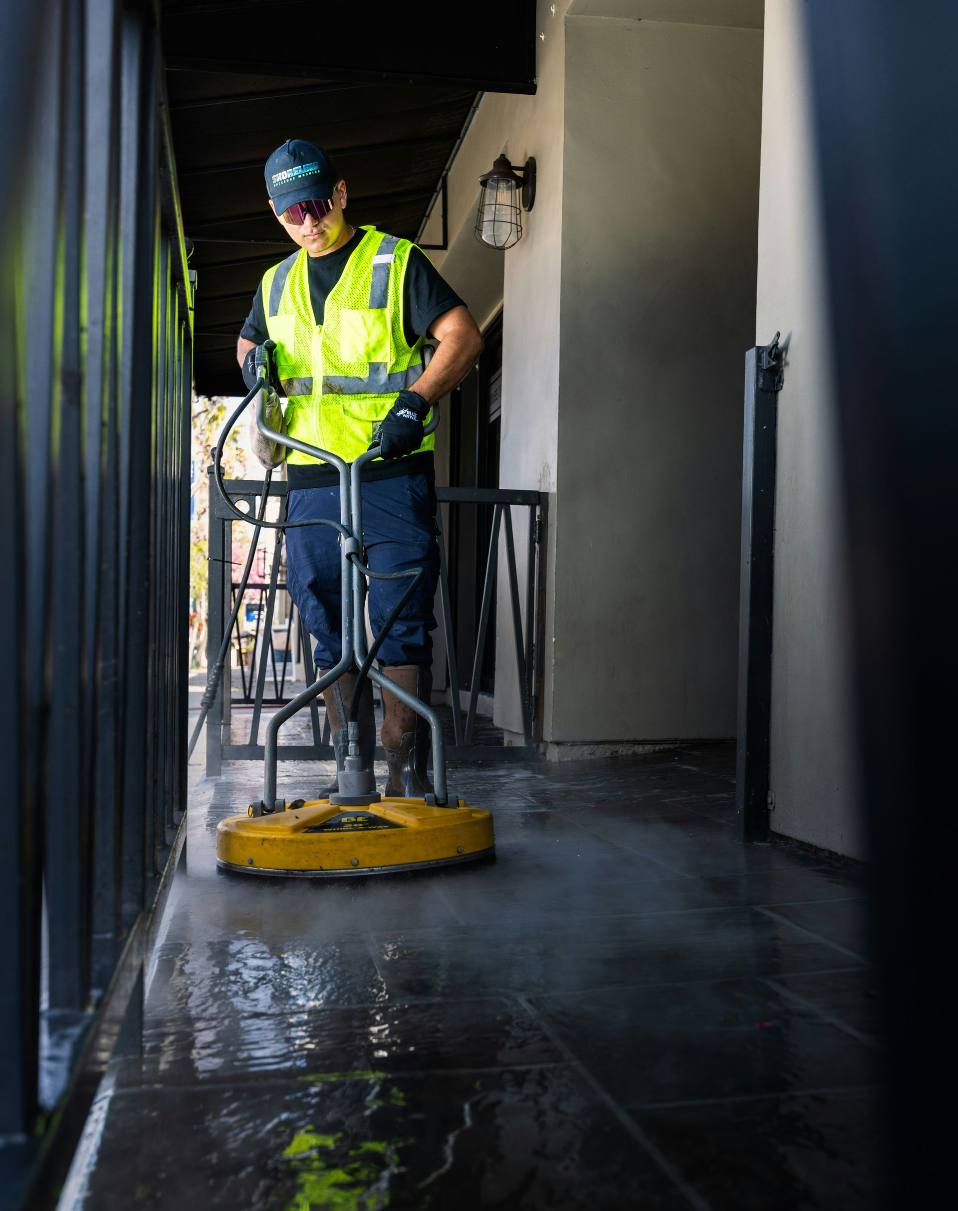 Person using a power washer to clean a dark stone walkway. They wear a safety vest and cap.