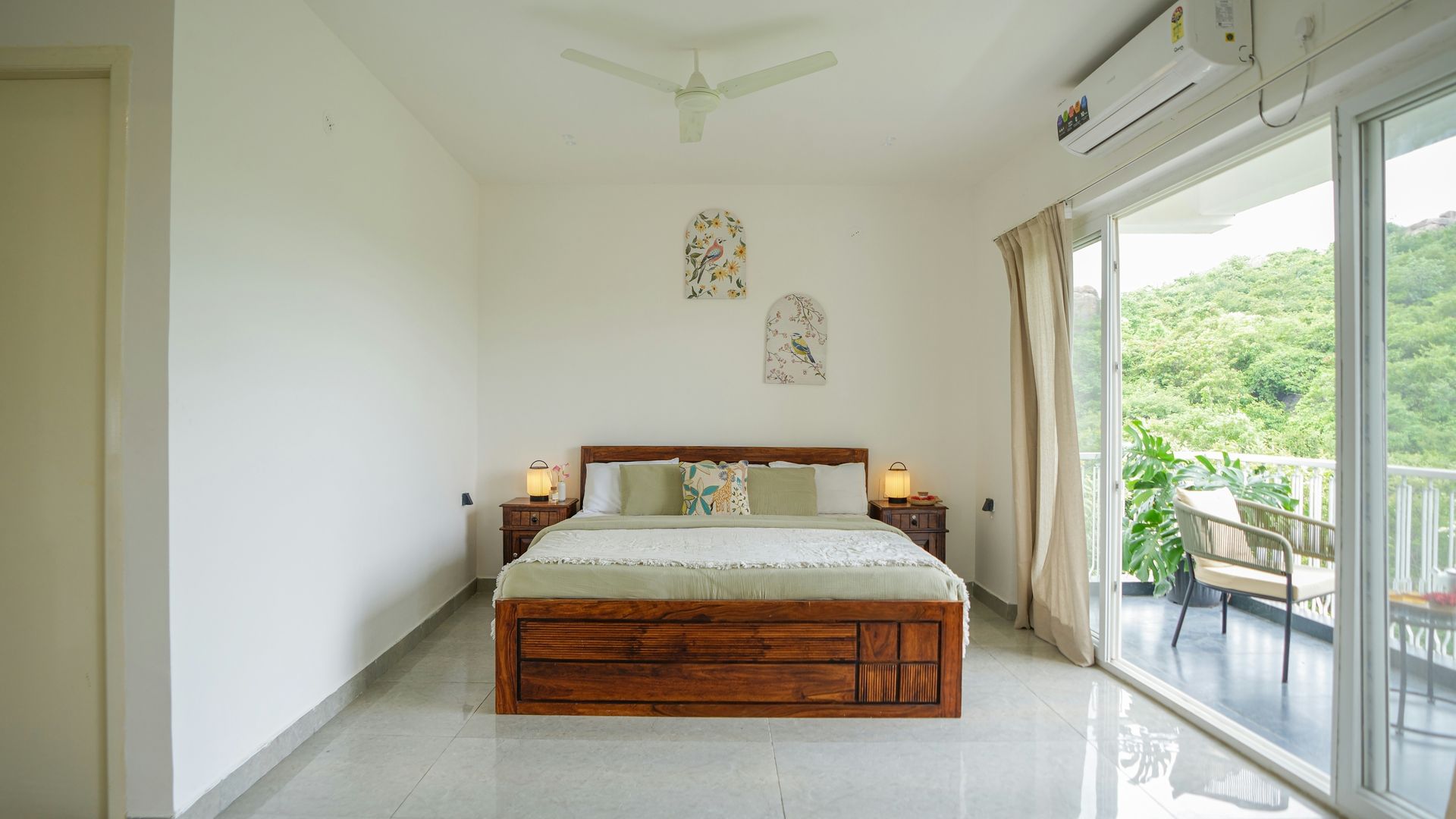 A modern bedroom with a wooden bed, white walls, decorative wall art, and a balcony view of lush green hills.