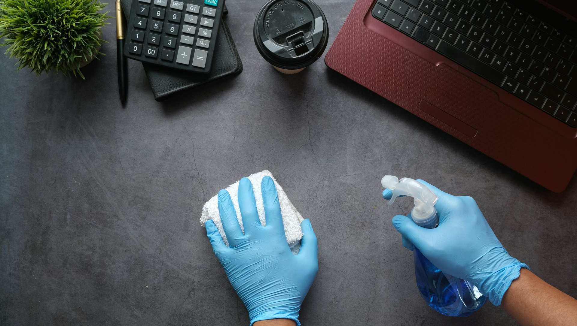 Hands in blue gloves sanitizing a dark desk with cleaning spray and cloth. Office items nearby.
