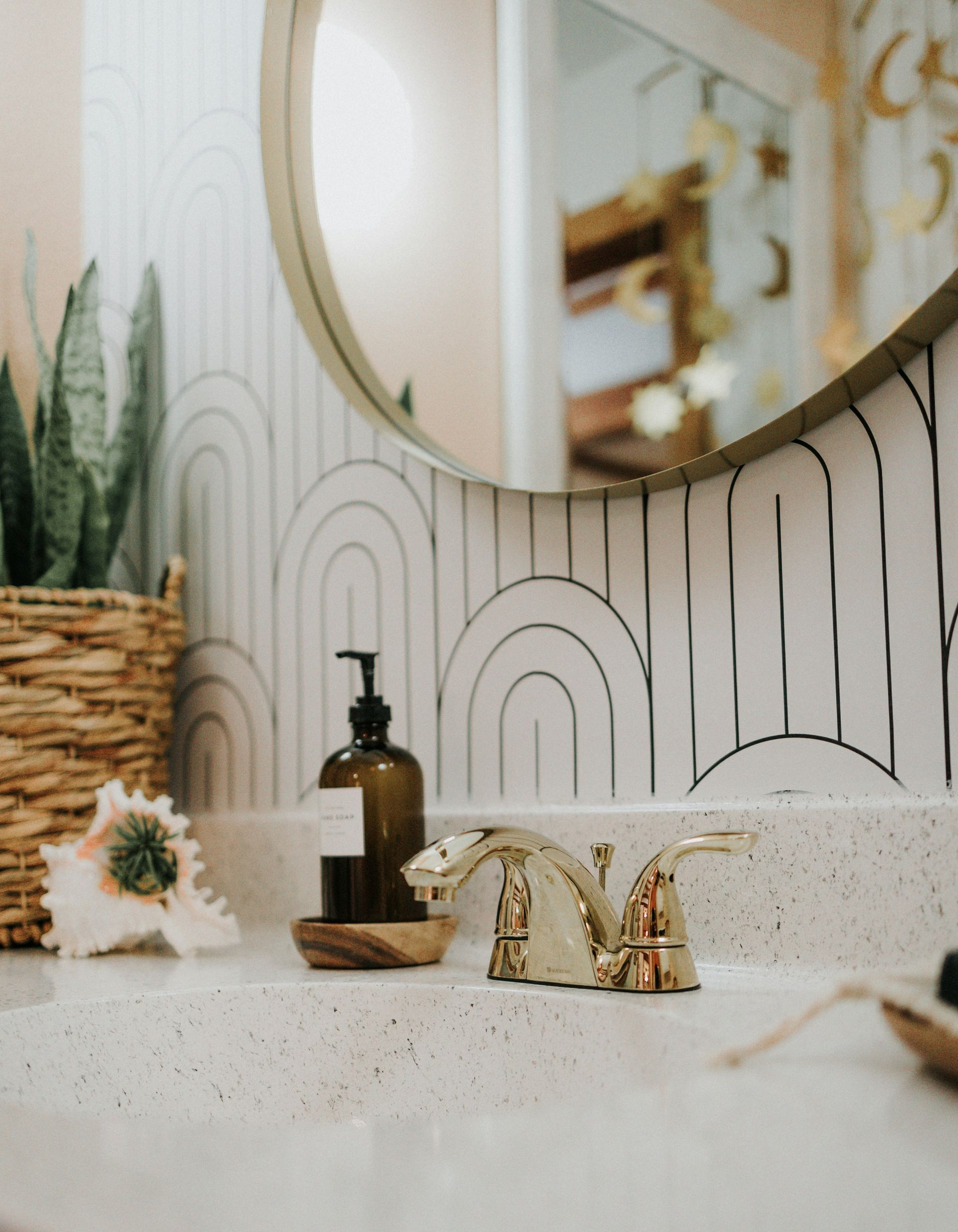 A bathroom vanity with a speckled countertop, gold faucet, soap dispenser, and houseplant against an arched-pattern wall.