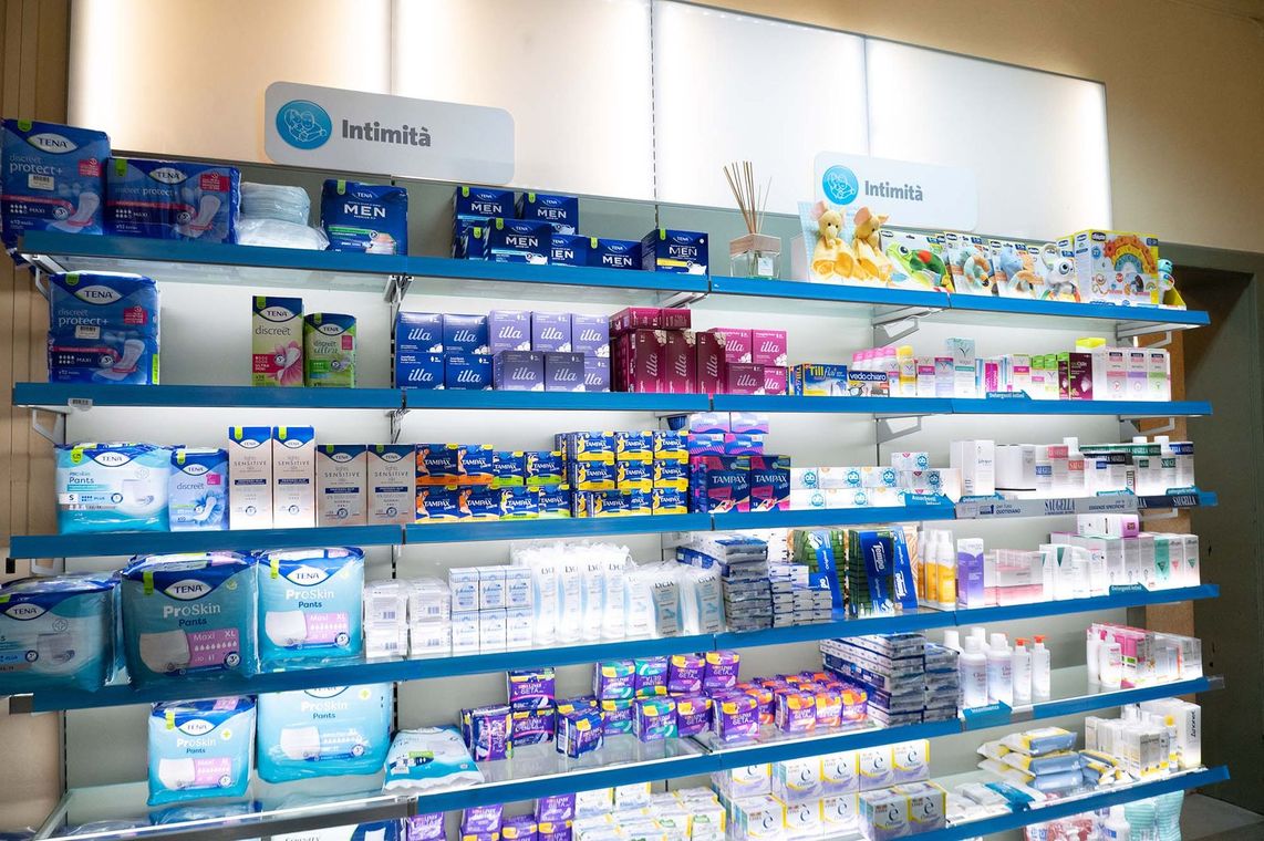 Una persona in camice bianco, con le braccia incrociate, tiene in mano uno stetoscopio, sullo sfondo sfocato di una farmacia.