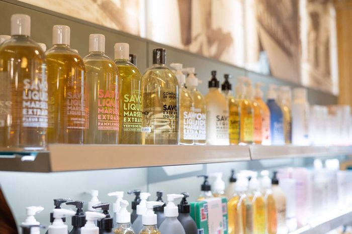 Una persona in camice bianco, con le braccia incrociate, tiene in mano uno stetoscopio, sullo sfondo sfocato di una farmacia.